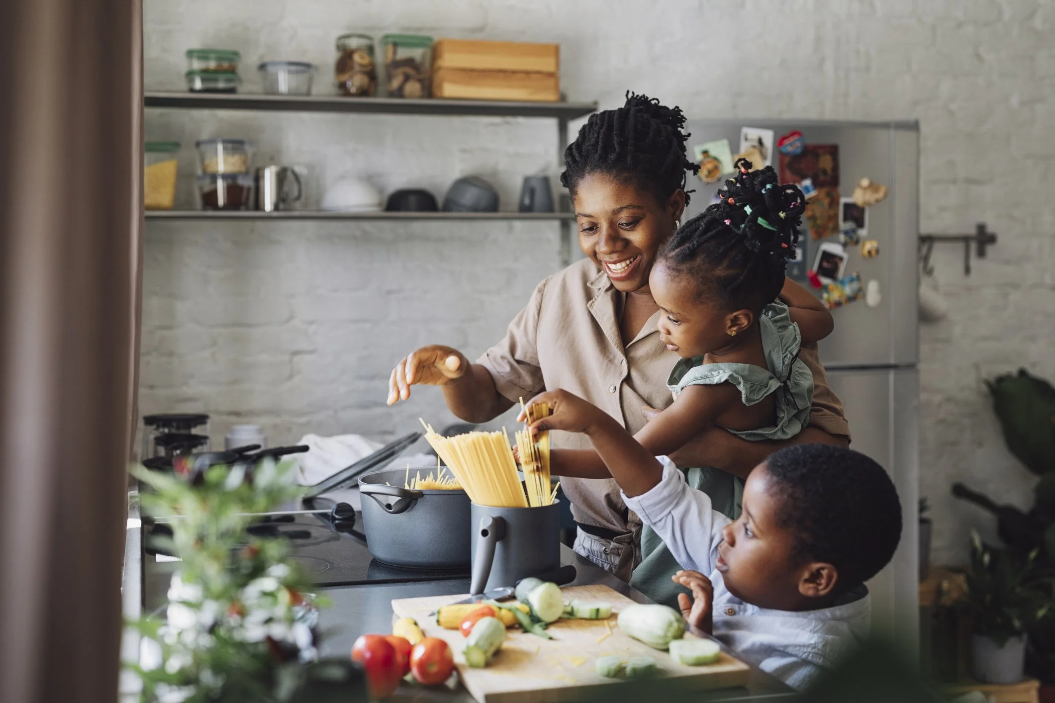 A woman with two children cooking in a modern kitchen, with vegetables and pasta on the counter.