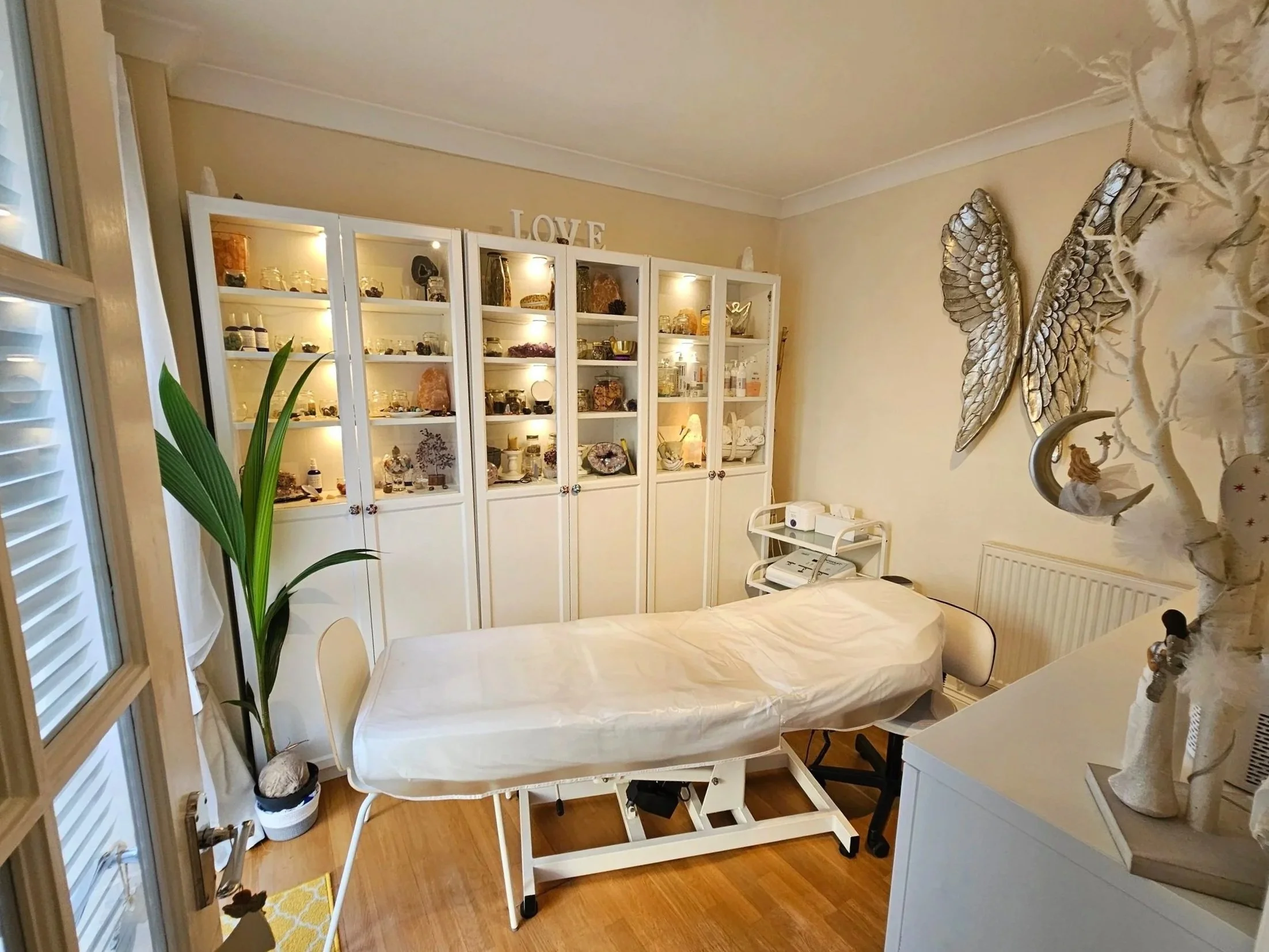 A cozy treatment room with a massage or therapy bed covered with a white sheet. To the right, there is a small white cart with medical or massage equipment. Behind the bed, a white cabinet displays various decorative items, crystals, and ornaments. A large potted plant with broad green leaves is near the window. The wall on the right has silver angel wing wall decor and a crescent moon with a small angel figure. The room has warm lighting creating a relaxing atmosphere.