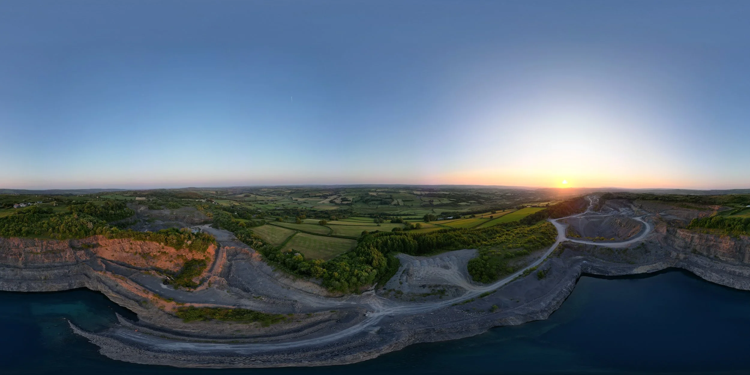 A panoramic aerial view of a landscape showing a large quarry near a body of water, with green fields, trees, and a sunset in the background.