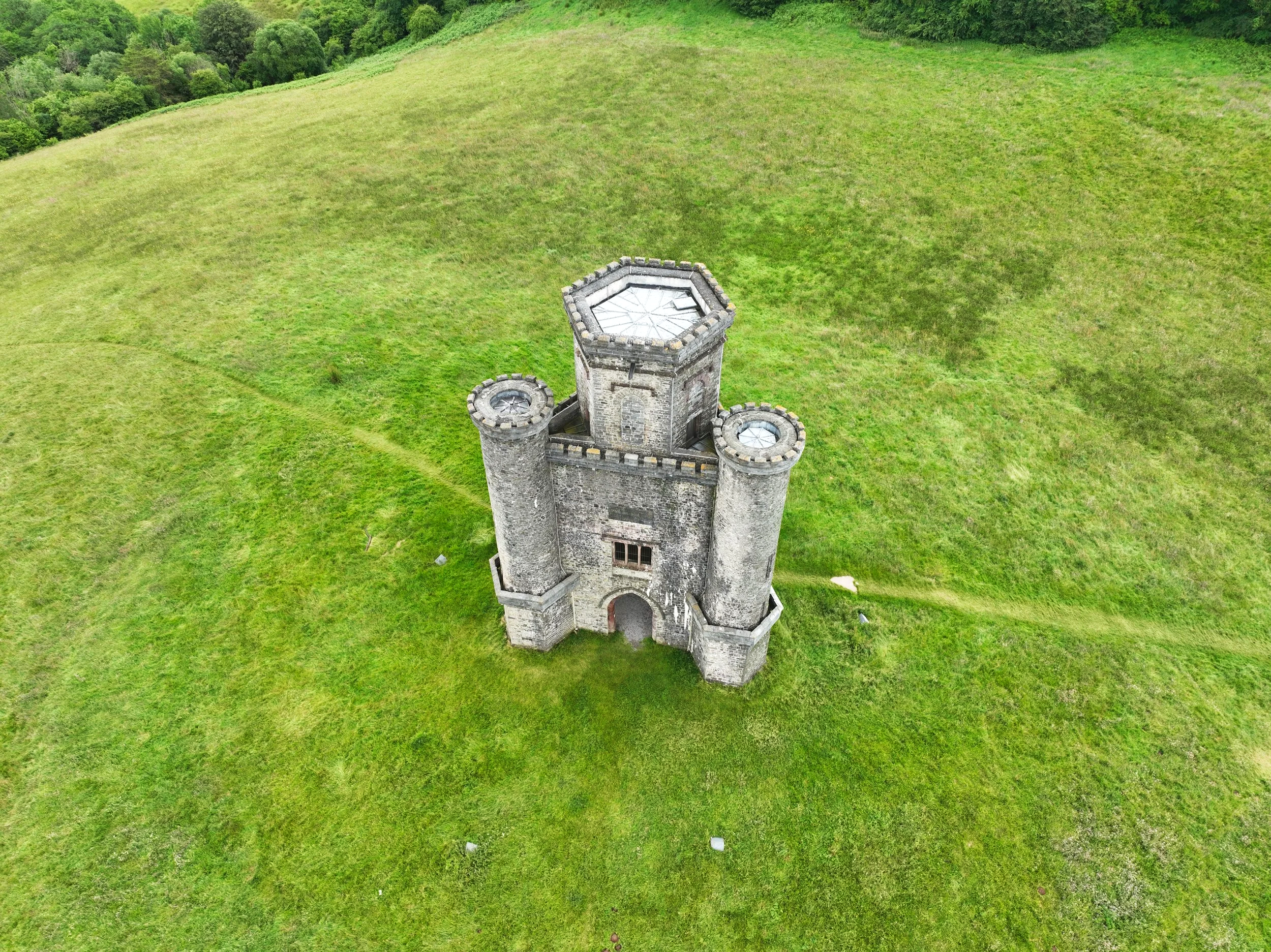 An aerial view of a small, ancient stone castle with three towers, situated on a grassy hill surrounded by lush green trees.