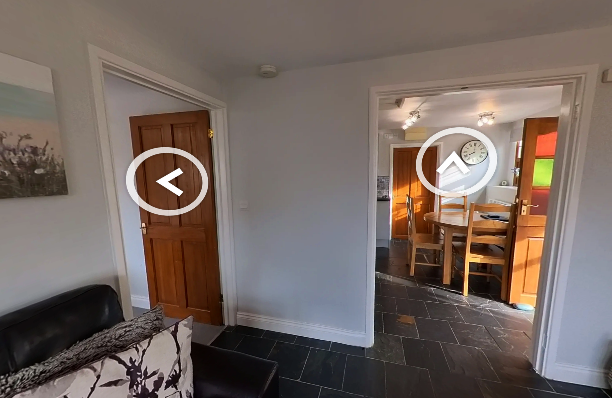 Living room with a door on the left and an open doorway leading to a dining area with a round table and chairs, dark slate flooring, and a wall clock.