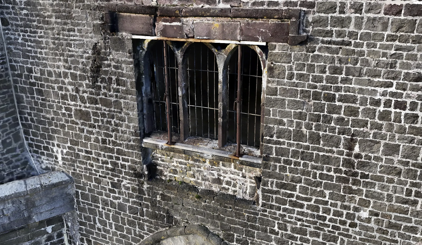 Close-up of a brick wall with a small arched window featuring metal bars and a wooden frame, some moss and grime on the bricks.