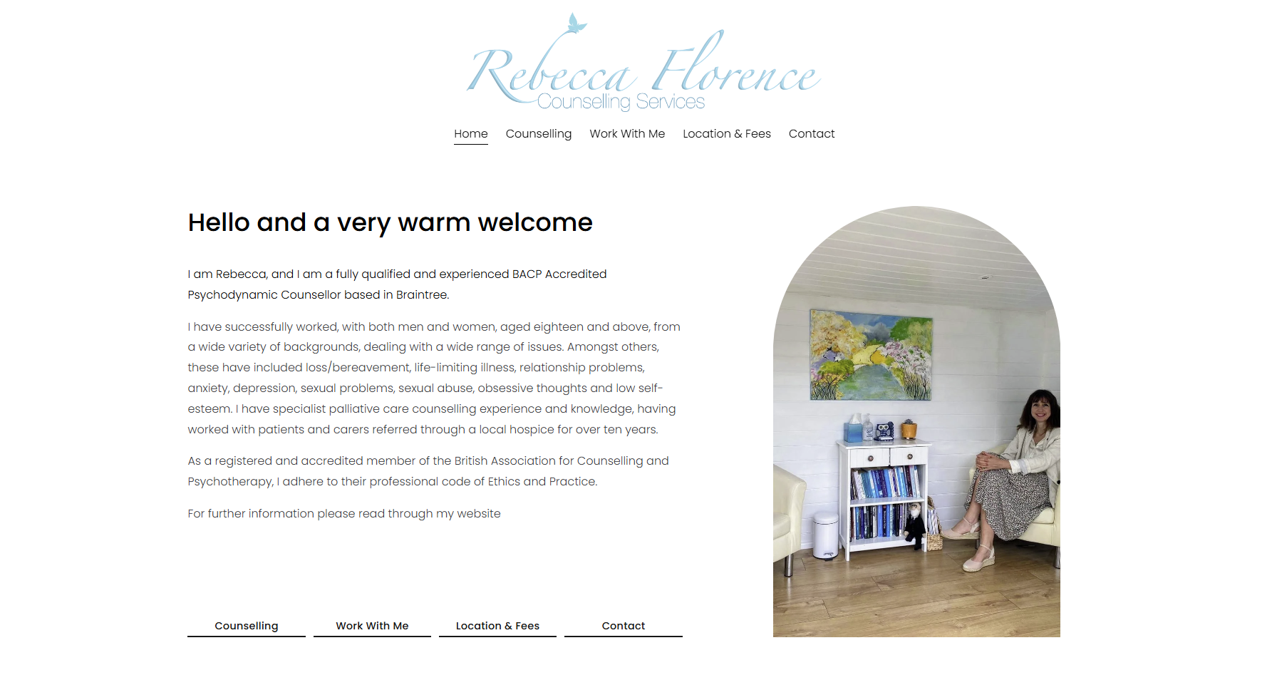 A woman sitting on a light-colored armchair in a bright, cozy room with white walls and wooden flooring, smiling at the camera. A colorful painting hangs on the wall behind her. To her left is a small white bookshelf filled with books, and a small item is placed on top of it. The room has a peaceful and welcoming atmosphere.