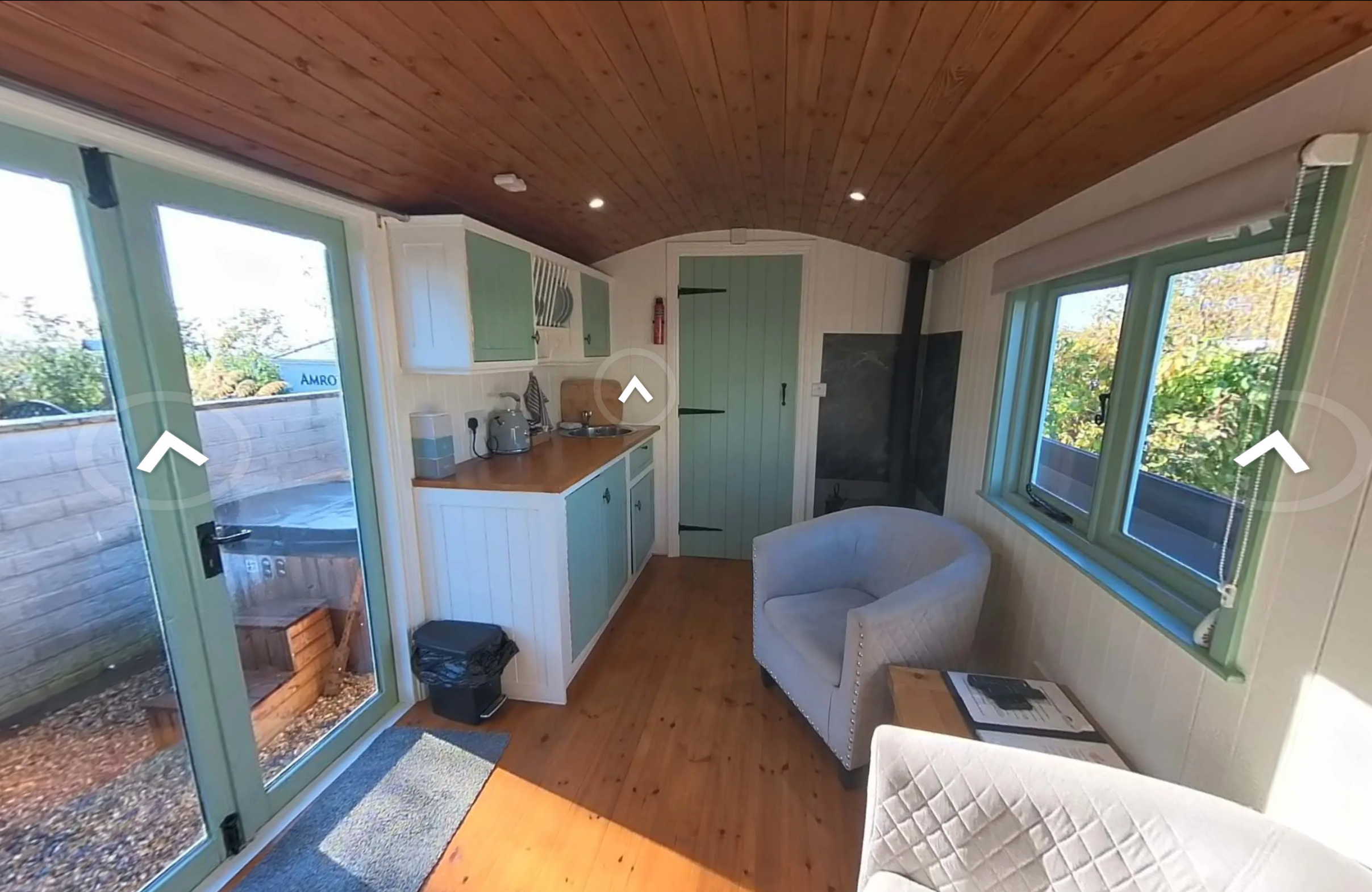 Interior of a small cozy kitchen with a wooden ceiling, green window and door frames, a small sitting area with a gray armchair, and a door leading outside.