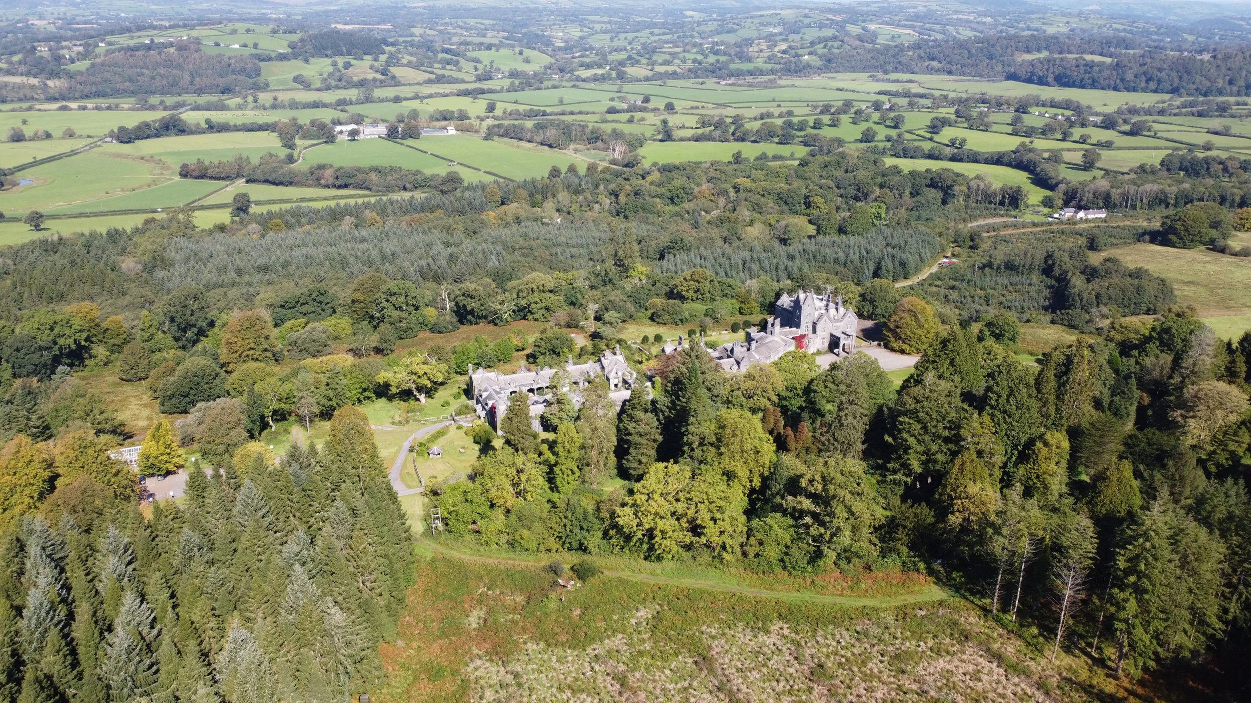 Aerial view of a large estate with a castle-like building surrounded by dense forest, open fields, and distant rolling hills.