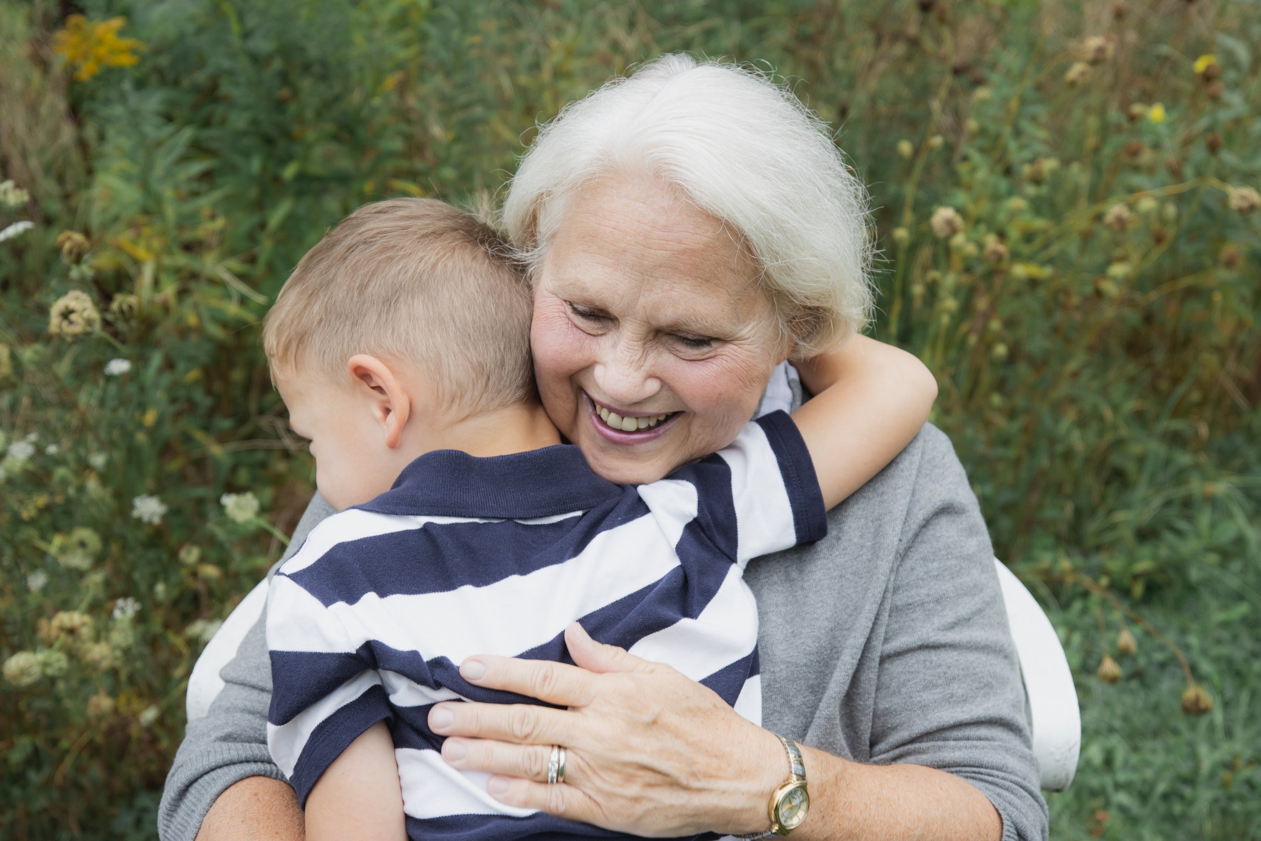 An elderly woman with white hair hugging a young boy with blonde hair outdoors, surrounded by green foliage and wildflowers.