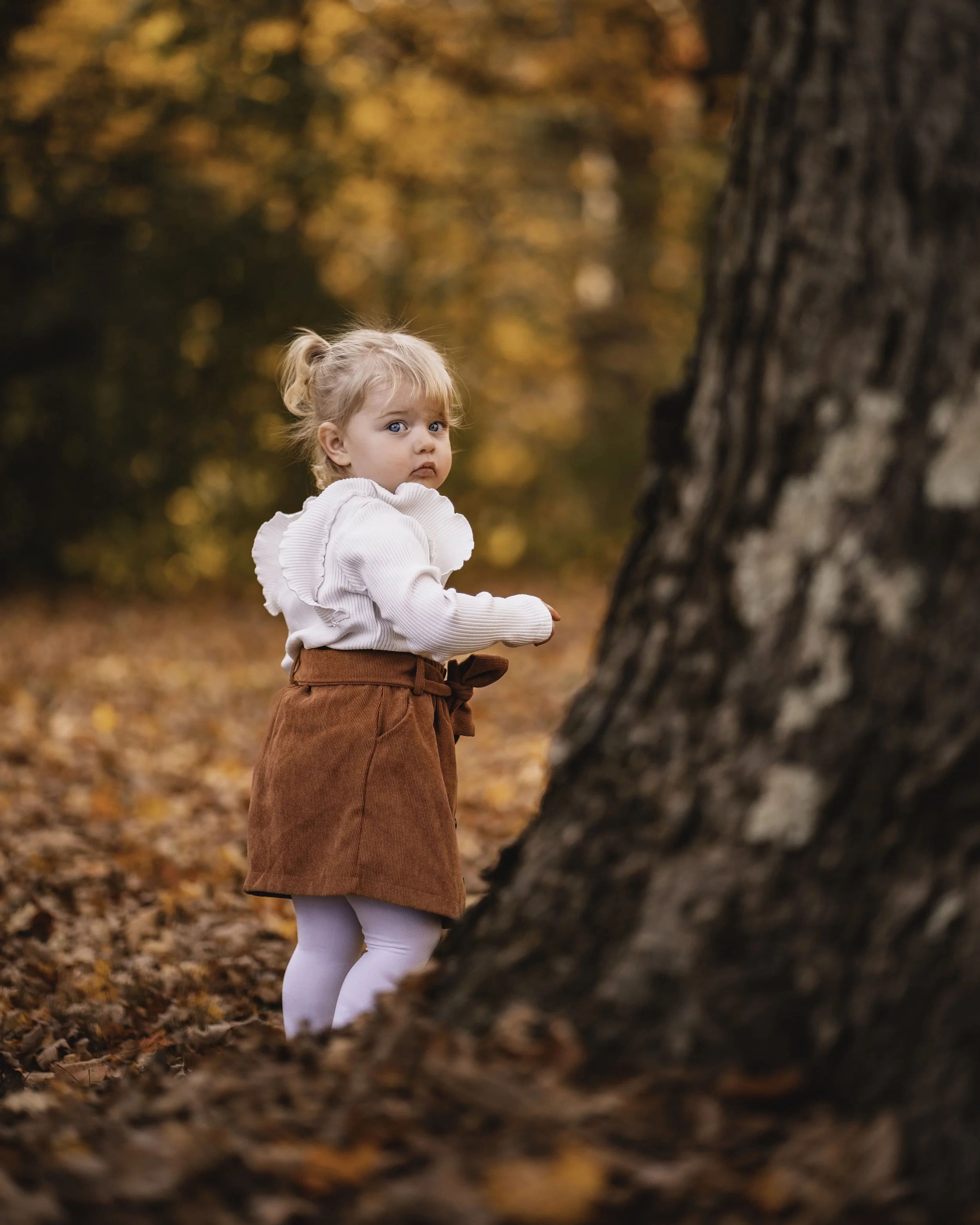 A young girl with blonde hair and blue eyes stands outdoors among autumn leaves, near a large tree trunk, wearing a white sweater, a rust-colored skirt, and white leggings, with a slightly cautious expression.