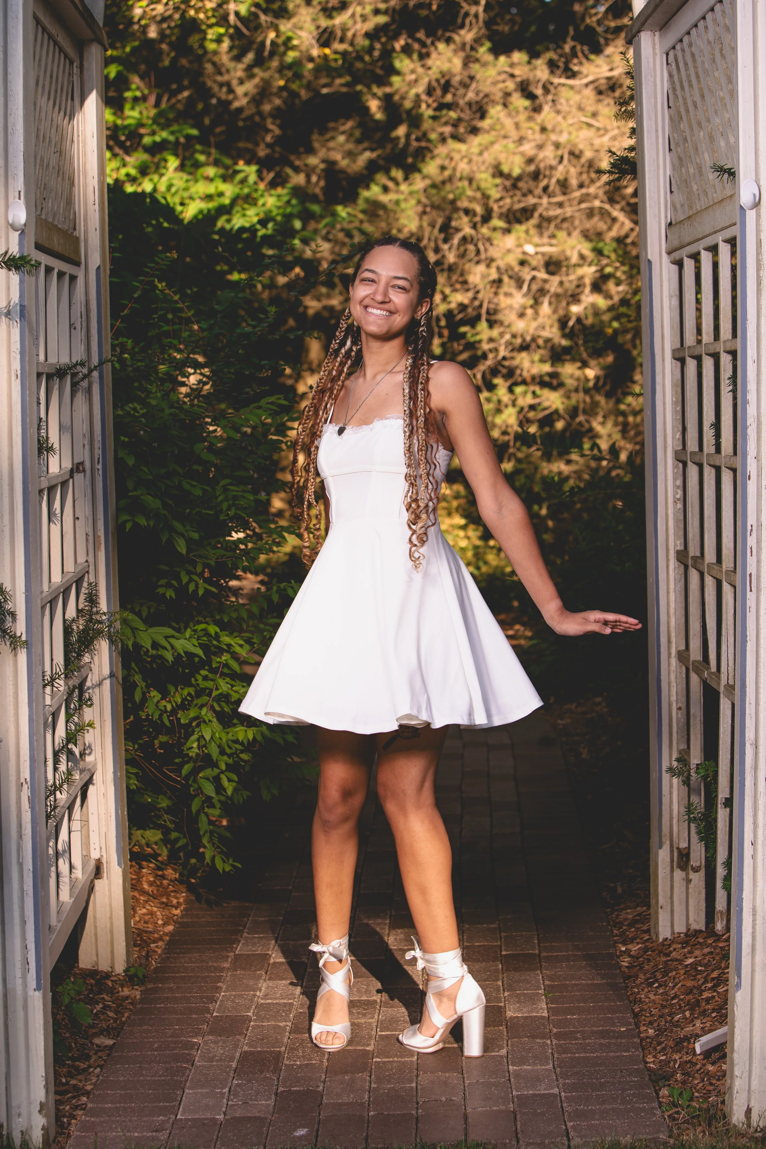 A woman in a white dress and white high heeled shoes with ankle ribbons standing in a garden gate, smiling.