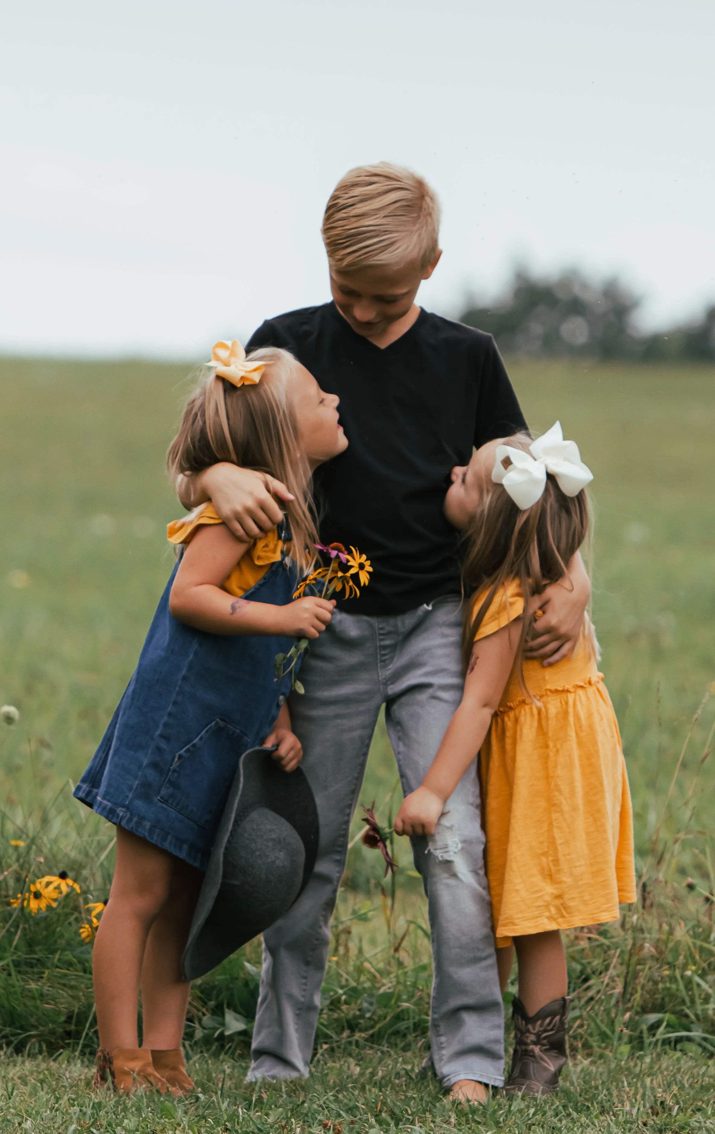 A boy with two young girls hugging outdoors in a grassy field, holding flowers.