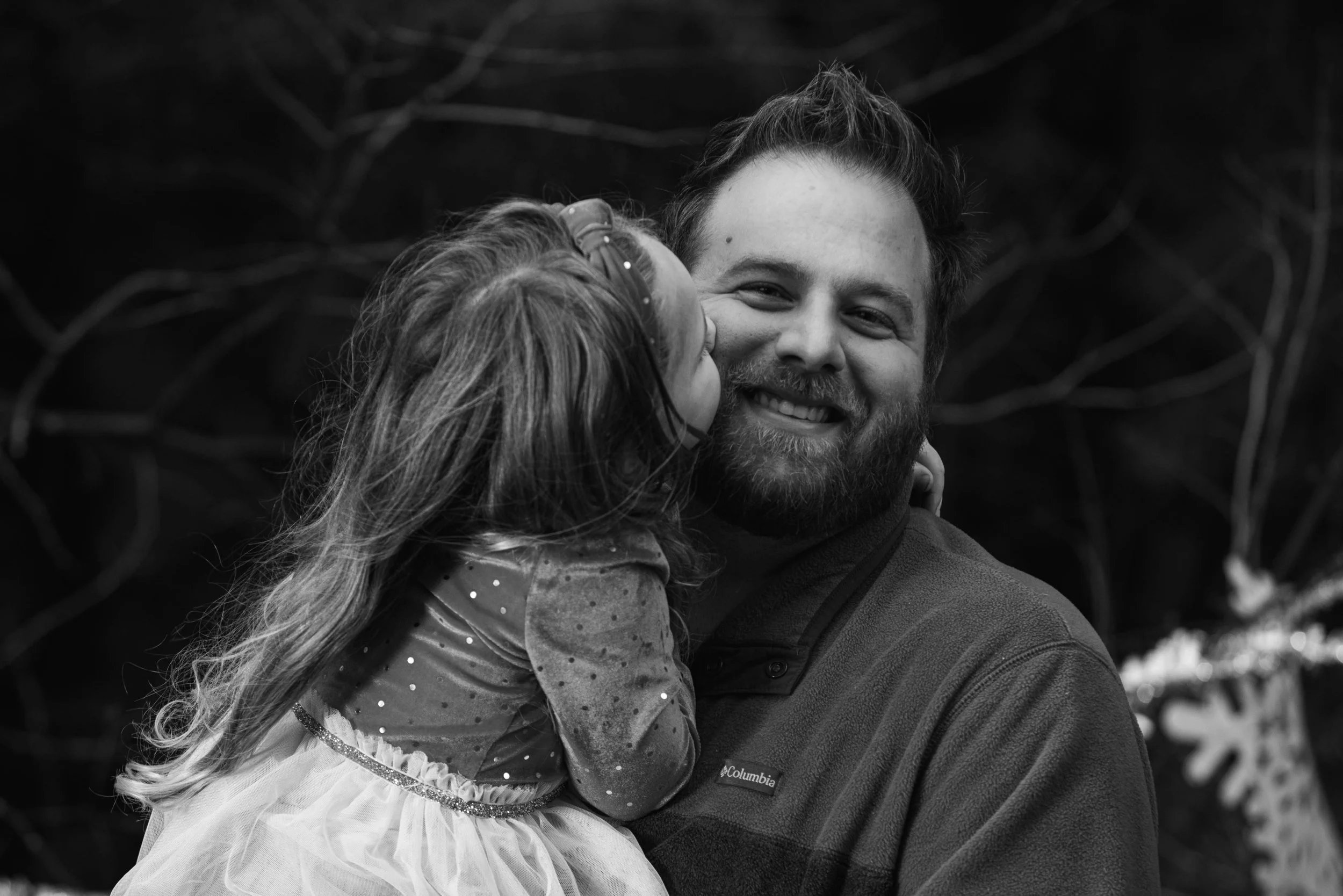 A man smiling while holding a young girl who is kissing his cheek in a black and white photo.