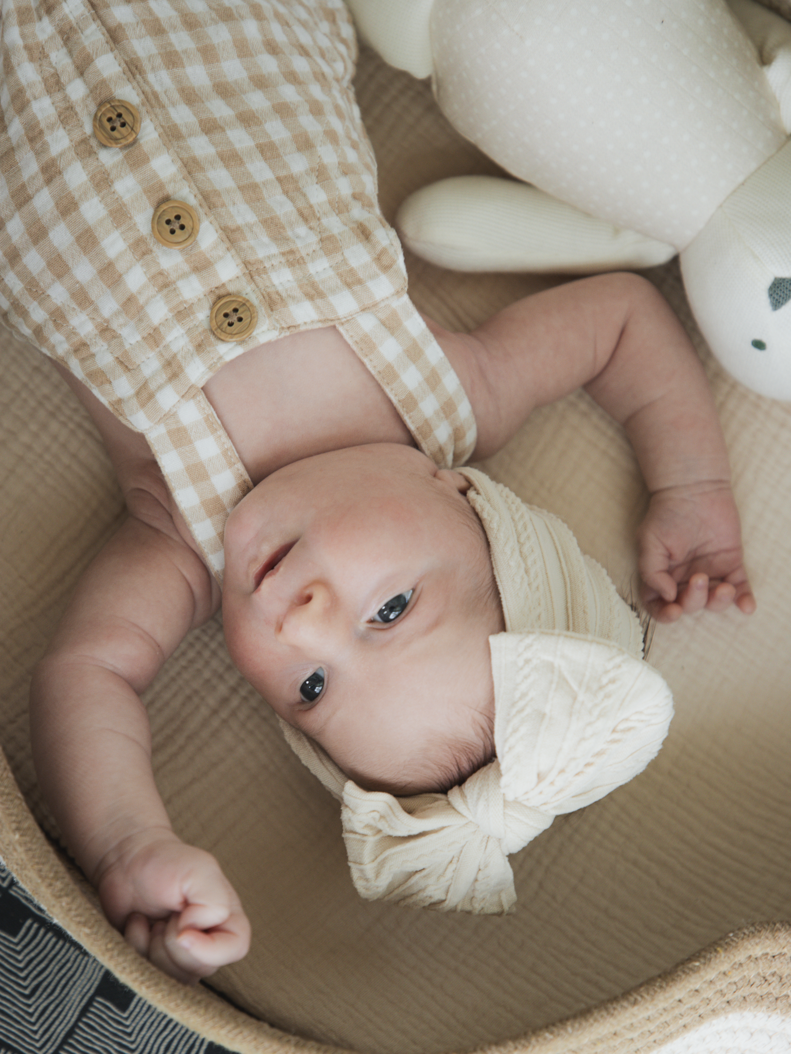 A baby lying on a beige changing pad, wearing a checked beige sleeveless top with buttons and a cream-colored knit hat with a bow, looking sideways with a curious expression.