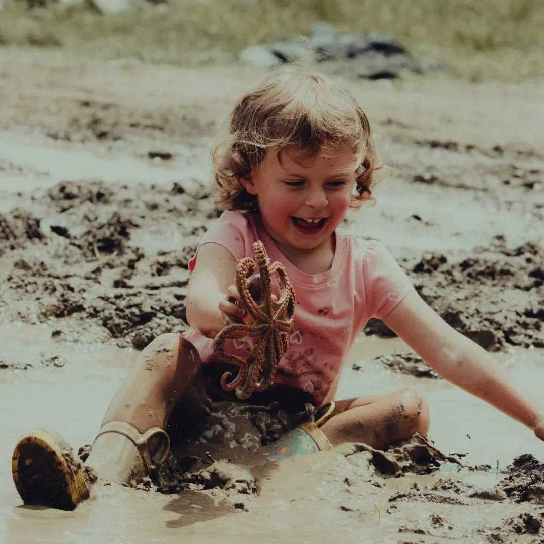 A young girl sitting in a muddy area, smiling and holding an octopus in her hand.