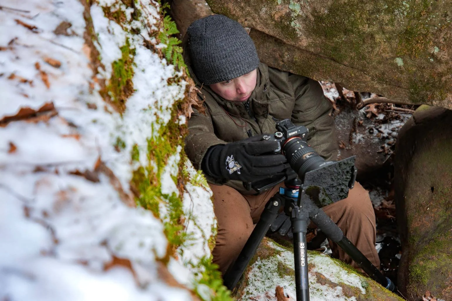 A person in winter clothing, including a gray knit hat and gloves, is taking a photograph with a camera on a tripod in a small, snowy, mossy, rocky crevice outdoors.
