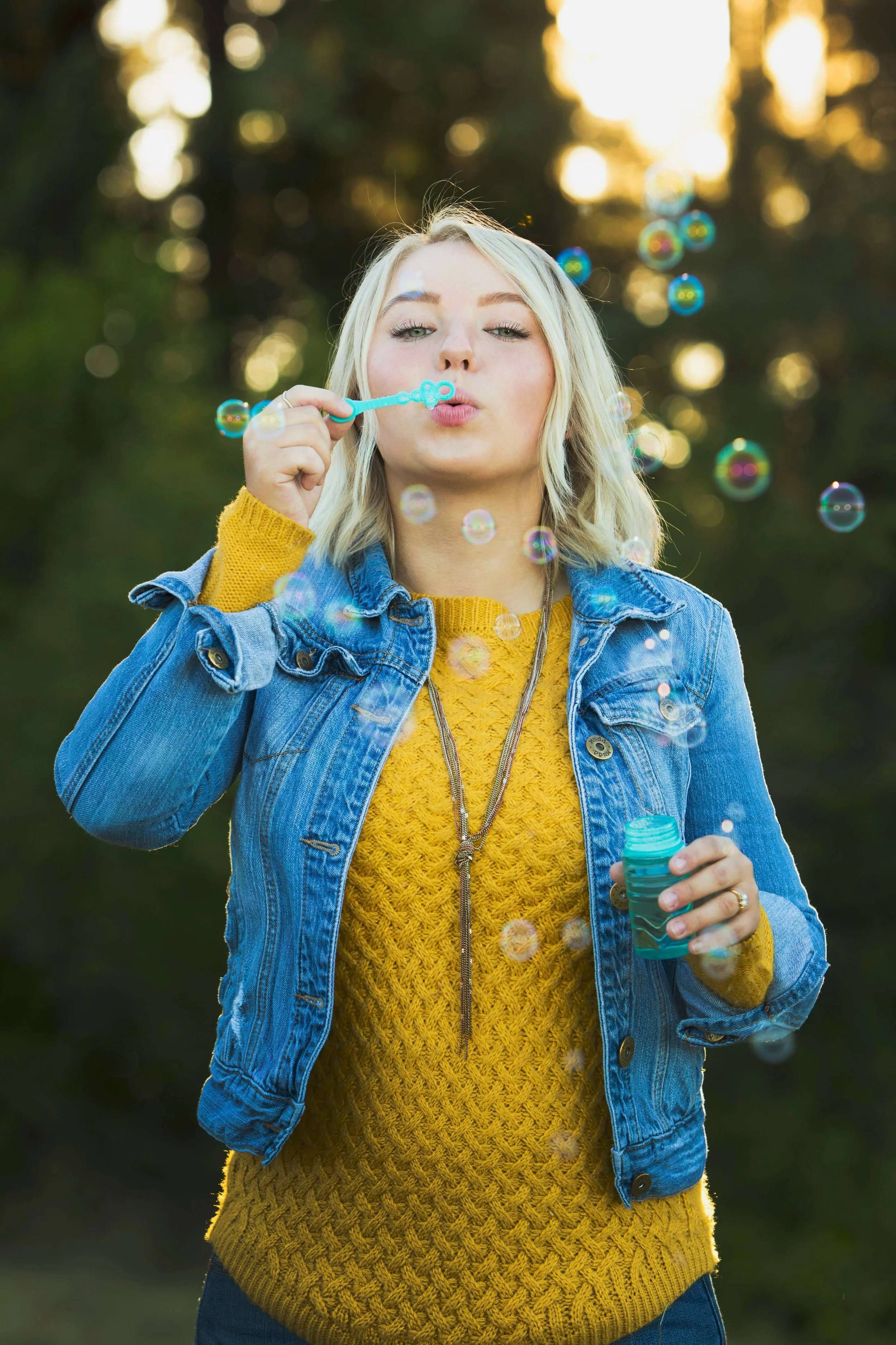 A young woman with blonde hair wearing a yellow sweater and blue denim jacket blowing bubbles outdoors with trees in the background.