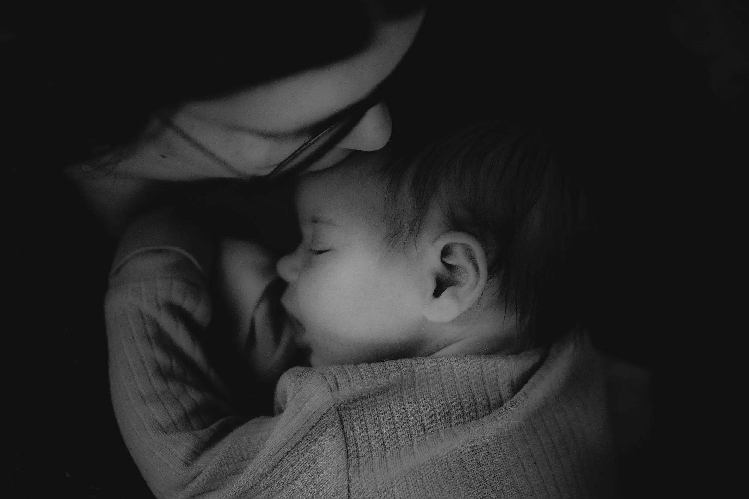 Black and white photo of a person with glasses gently kissing a young child's forehead while the child sleeps peacefully with eyes closed.