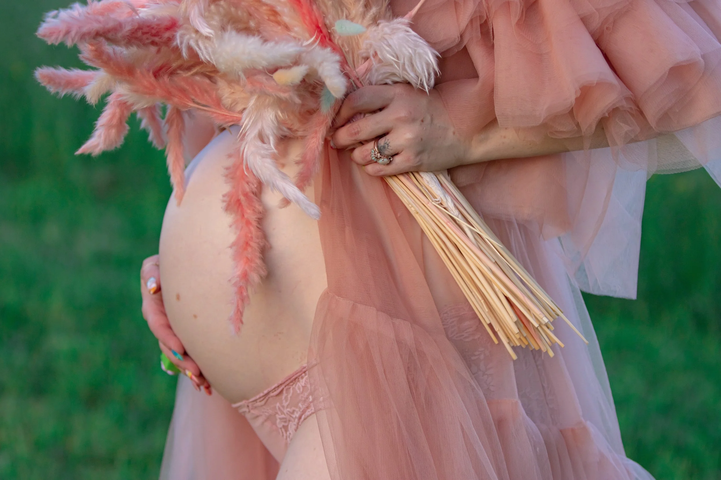 A person holding a bouquet of pink and white feathers, dressed in peach-colored, semi-transparent tulle fabric, with a hand adorned with rings and painted nails, in front of a green background.