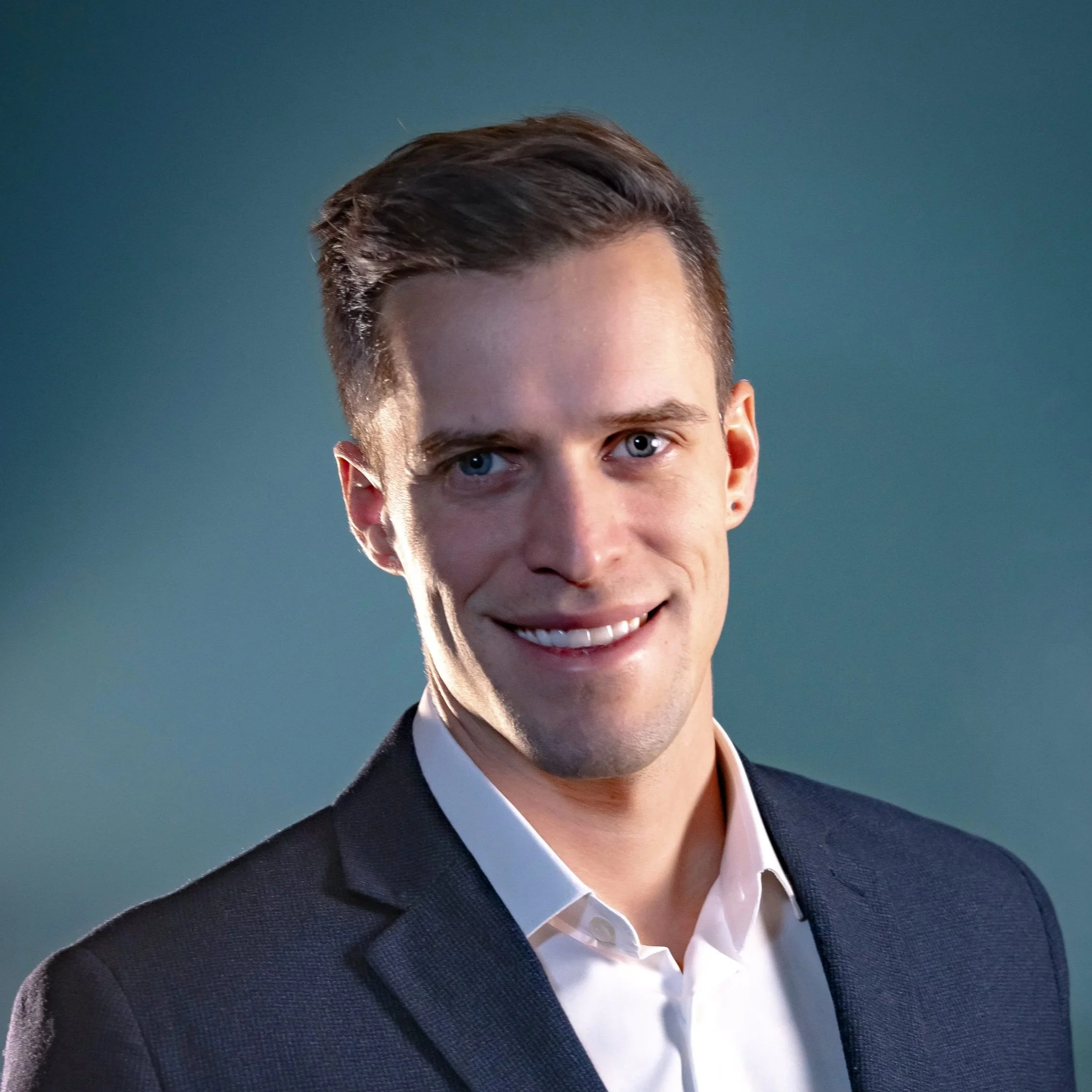 Close-up of a young man smiling, with blue eyes, wearing a dark blazer and white shirt, against a blue background.