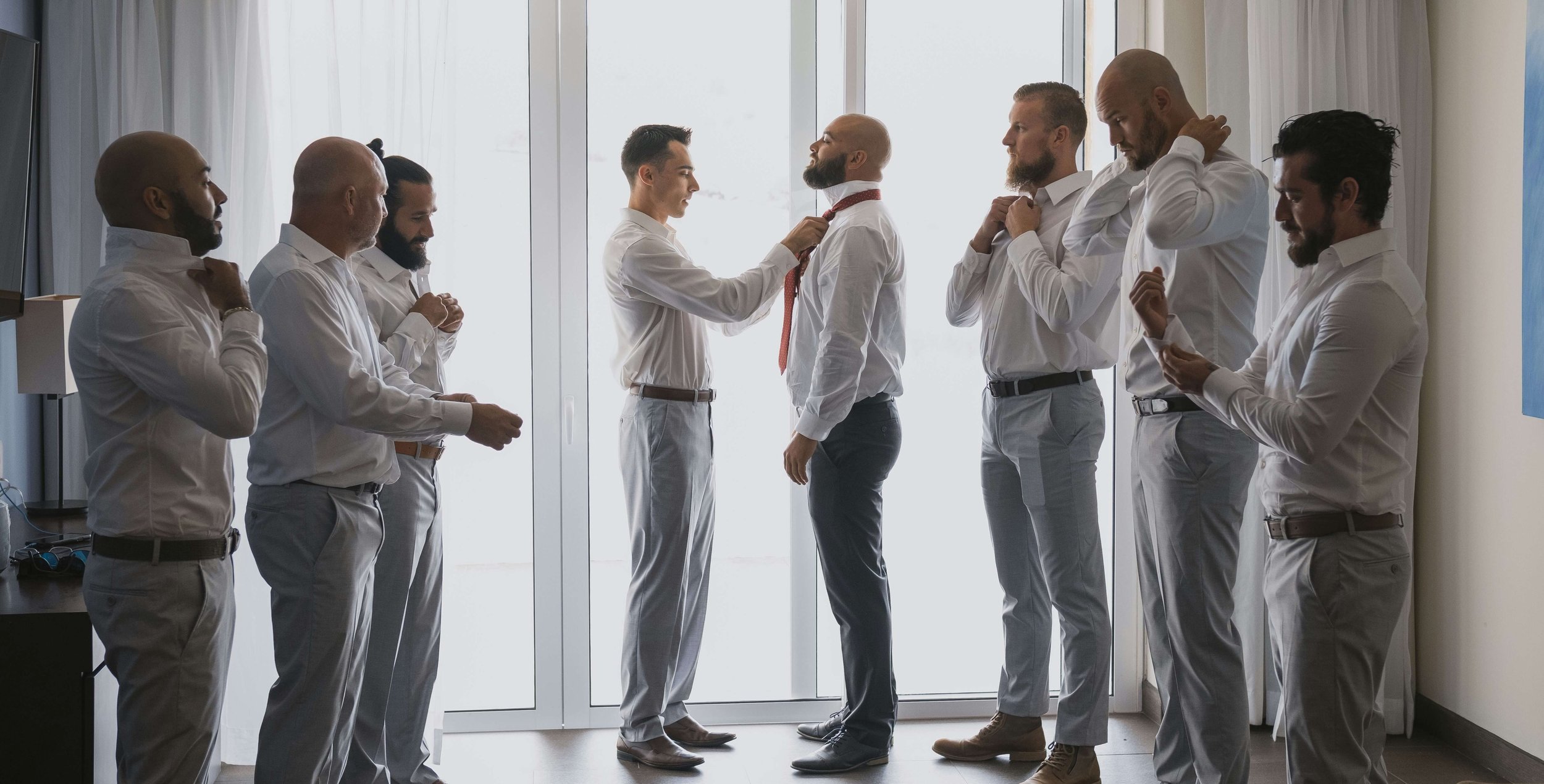 Group of men in formal attire, preparing for a team photo in a bright room with a large window, as one man helps another with a red tie.