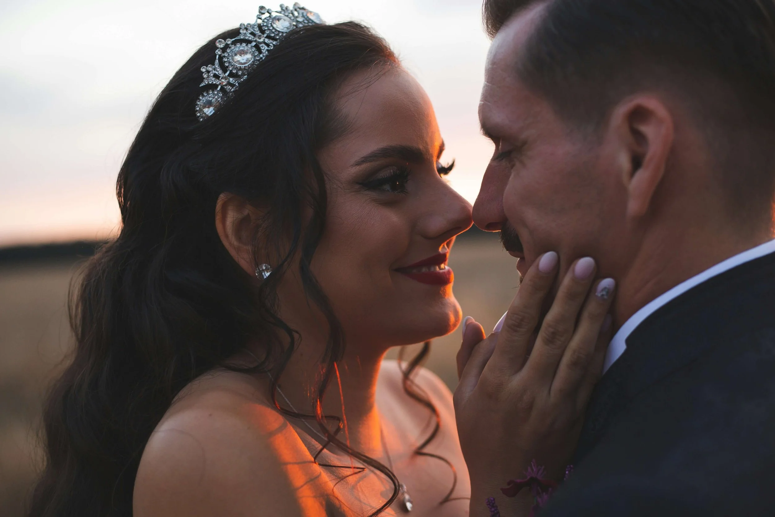 A couple is closely facing each other during sunset, with the woman gently touching the man's face, both smiling softly.