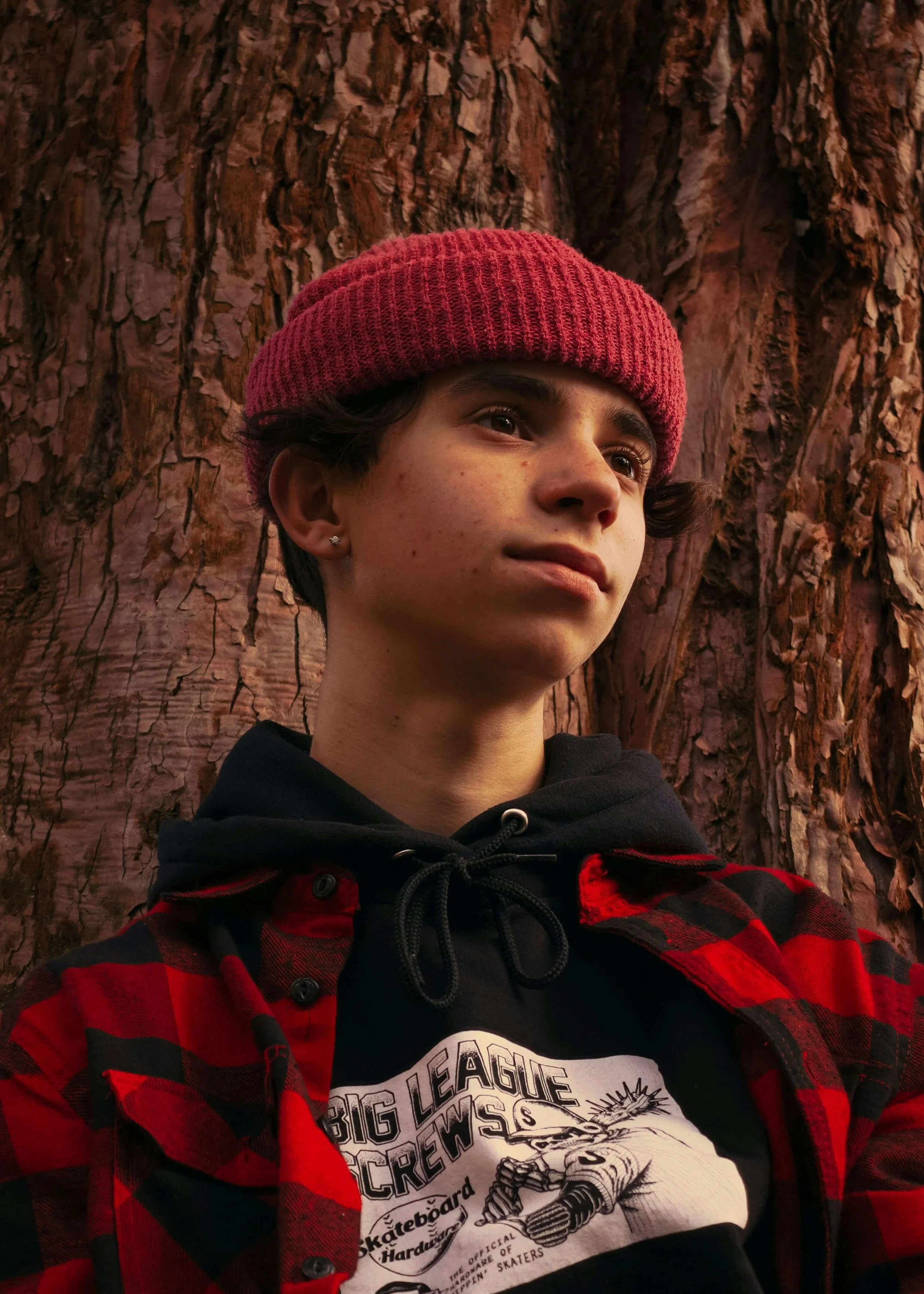 A young person with short dark hair, wearing a red knit beanie and a black hoodie, stands in front of a large tree with textured bark, dressed in a red and black plaid jacket.