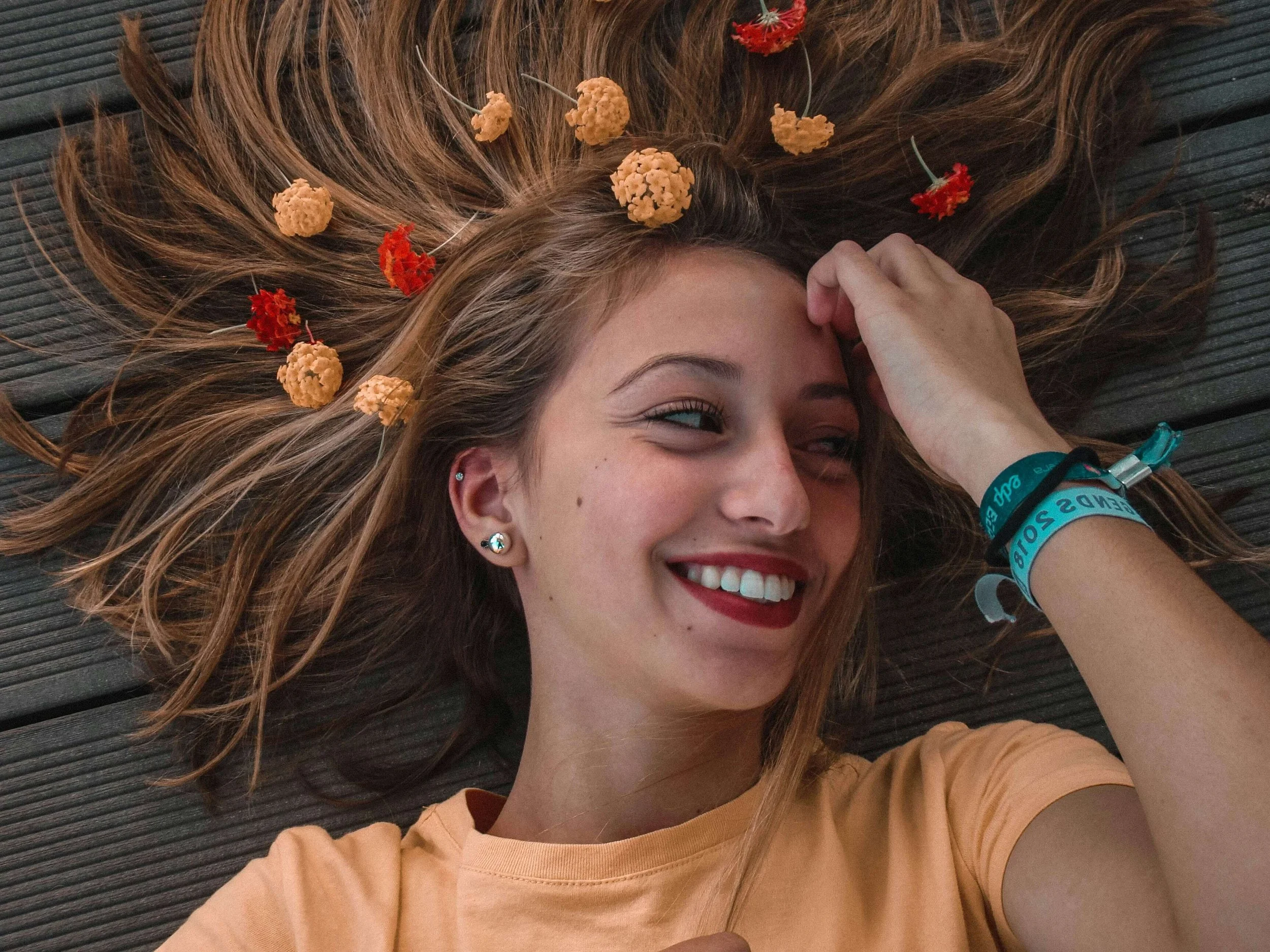 A smiling young woman with long, wavy hair lying on a wooden surface, surrounded by small brown and red flowers placed in her hair.