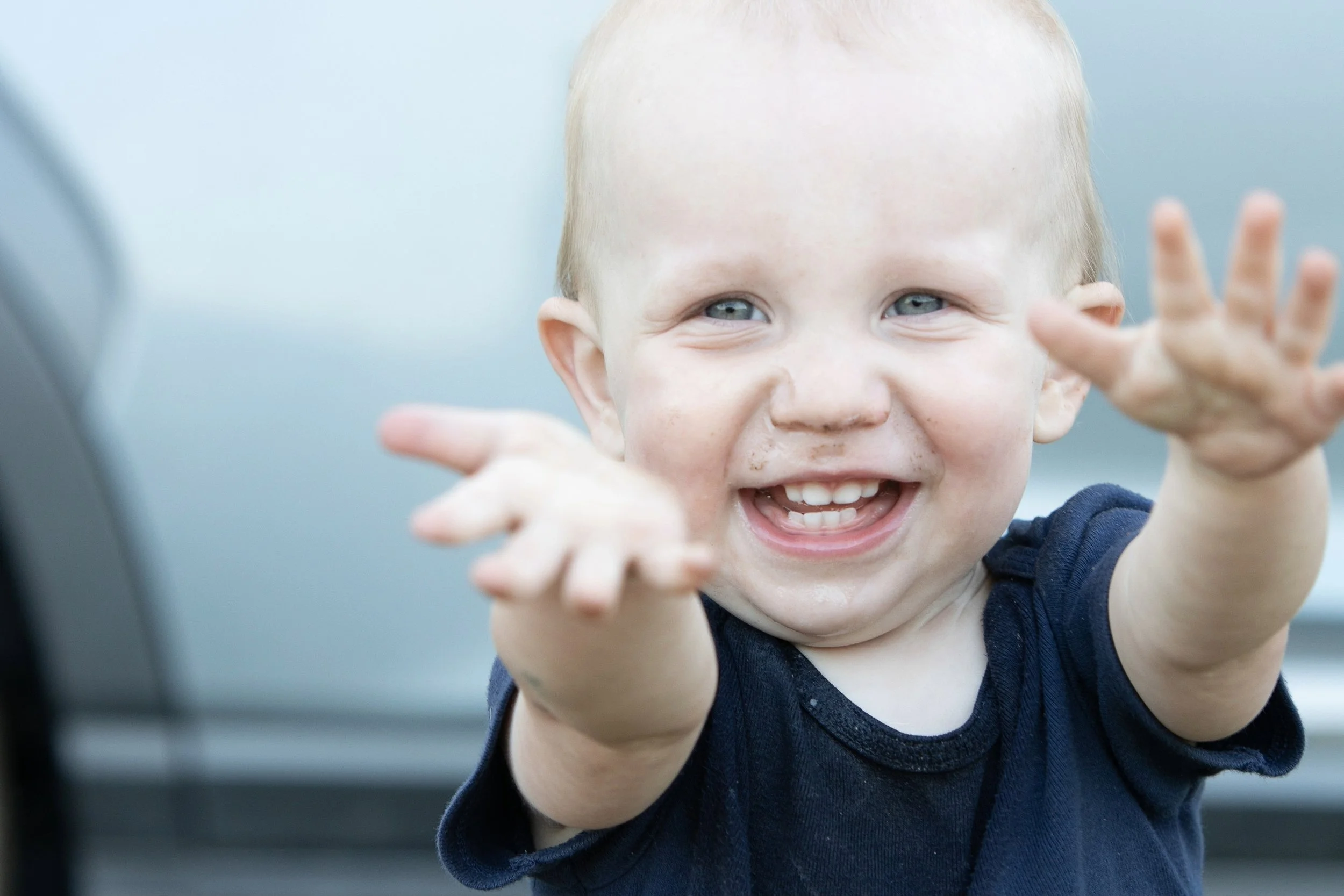 Close-up of a happy young boy with blonde hair and blue eyes reaching out with both hands toward the camera.