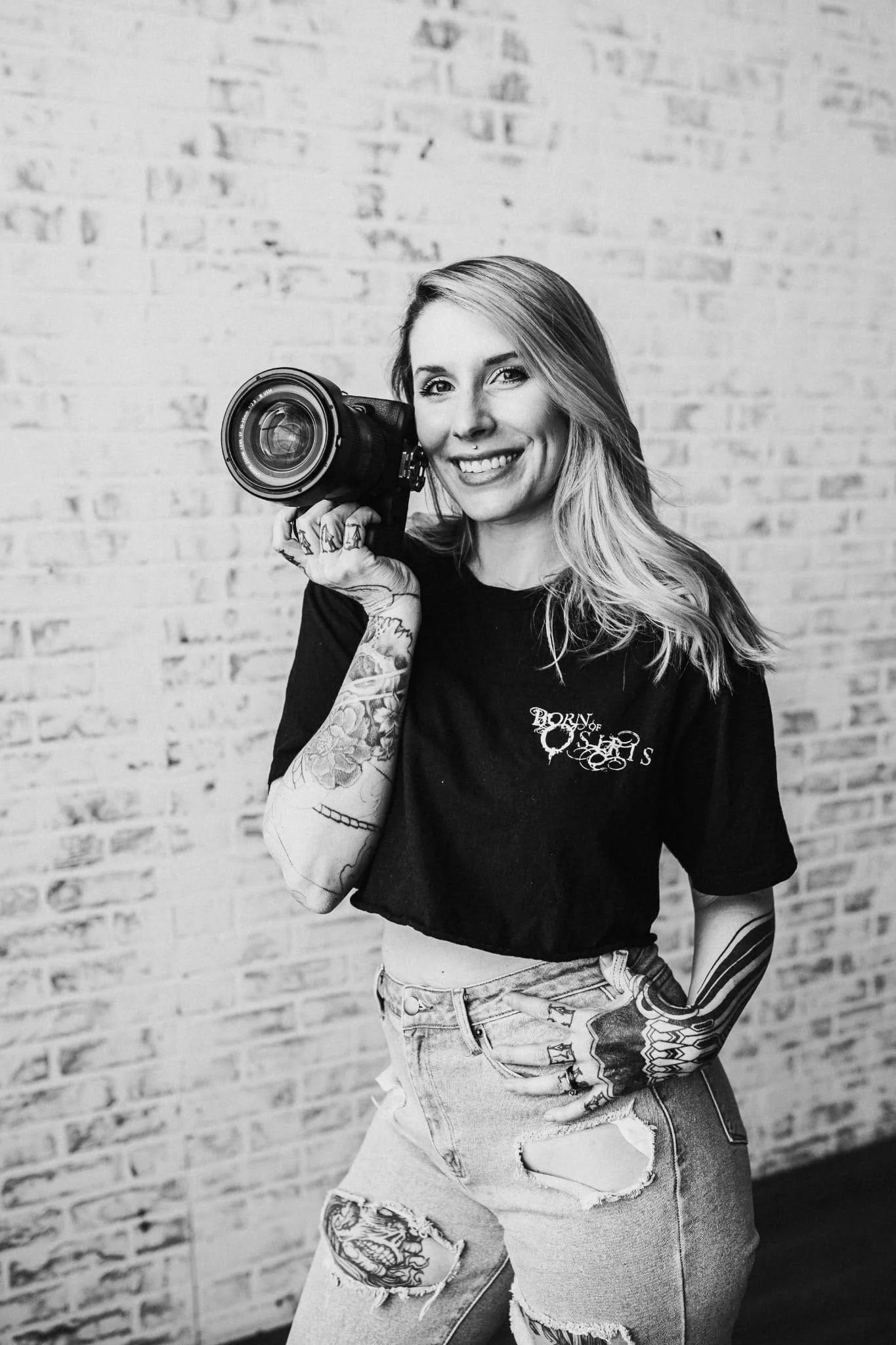 A young smiling woman with tattoos on her arm and leg, holding a professional camera close to her face in front of a brick wall.