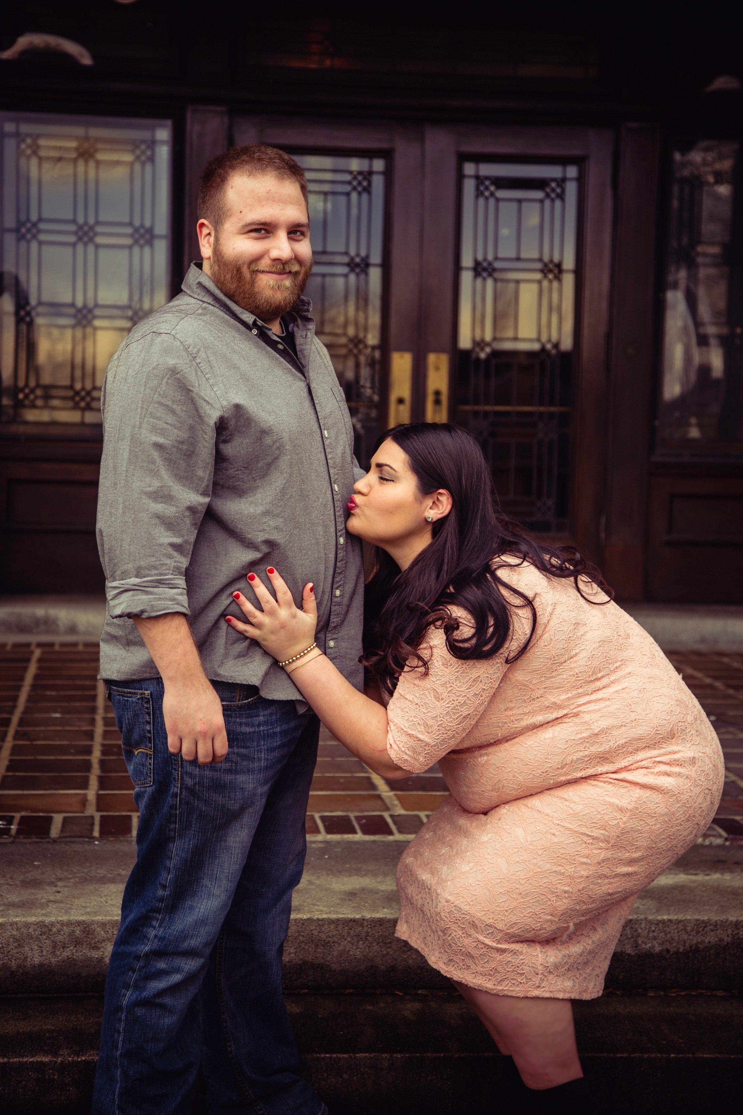 A man with a beard and a woman with long dark hair, dressed in a pink lace dress, sharing a tender moment outdoors in front of a dark wooden door with glass panels. The woman is kissing the man's chest, and her hand is on his stomach.