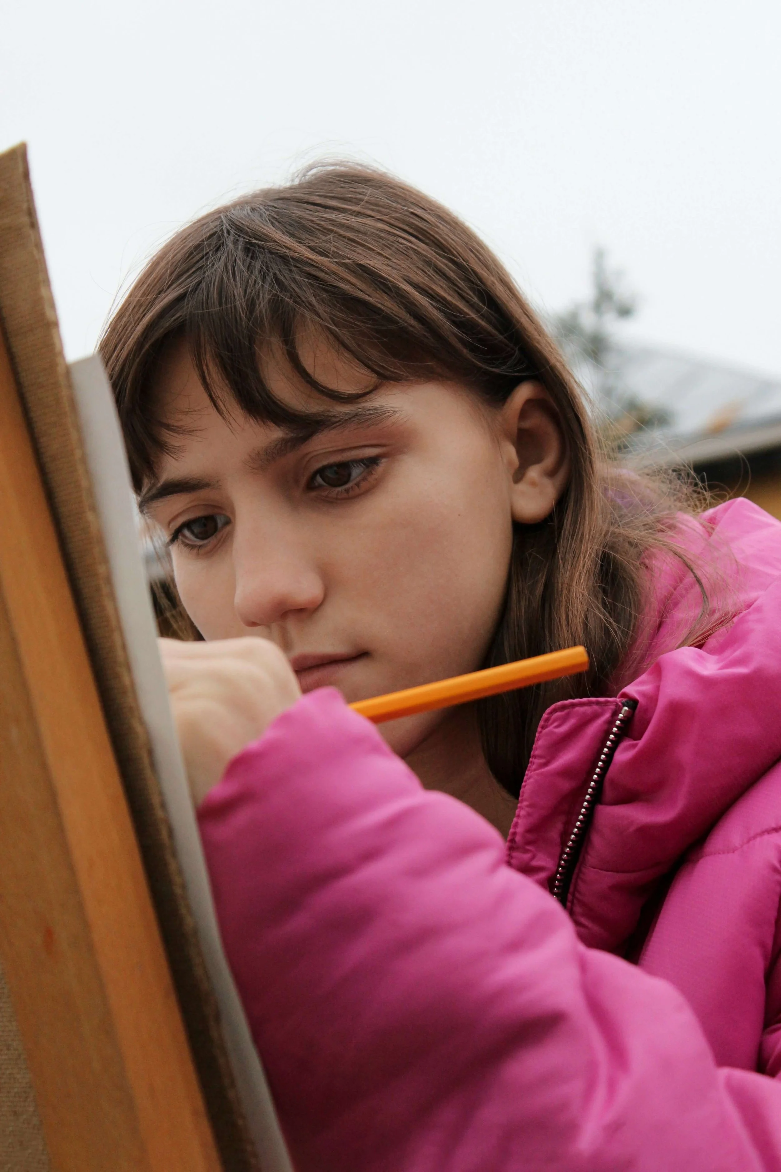 Young girl with brown hair and brown eyes wearing a pink jacket, sitting outdoors, and writing or drawing with an orange pencil on a notepad.