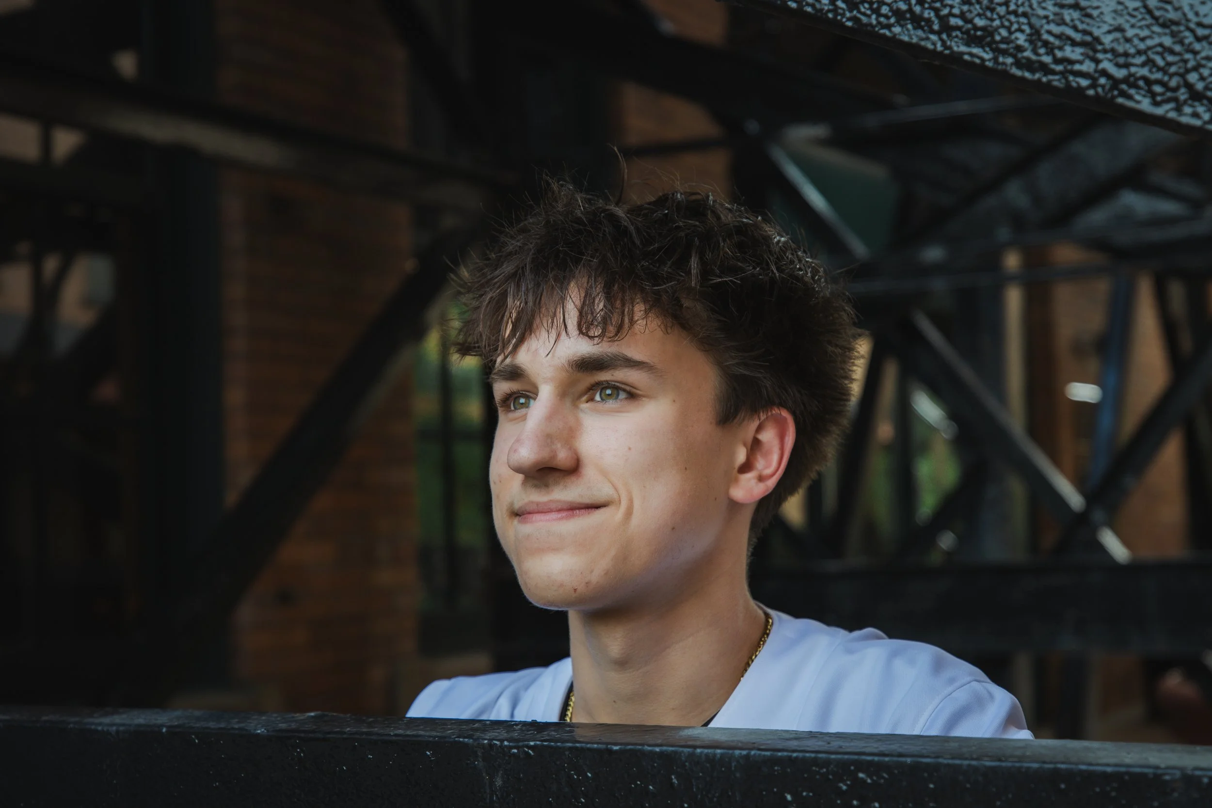 Close-up of a young man with curly brown hair and fair skin smiling and looking to the side, outdoors in front of a brick building and black metal structure.