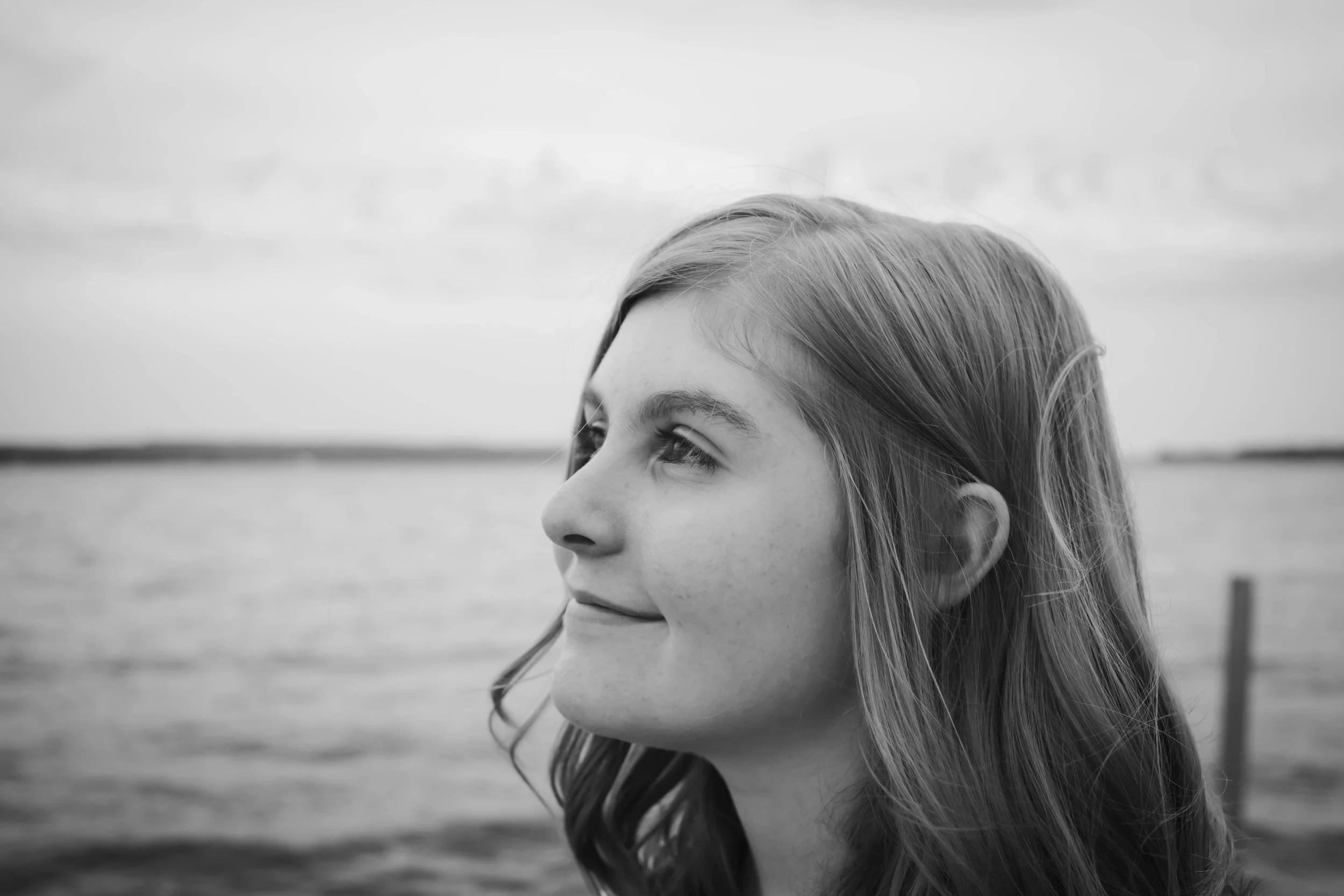 Black and white photo of a young woman with long hair looking to the left with a serene expression, standing outdoors by a body of water with a cloudy sky.