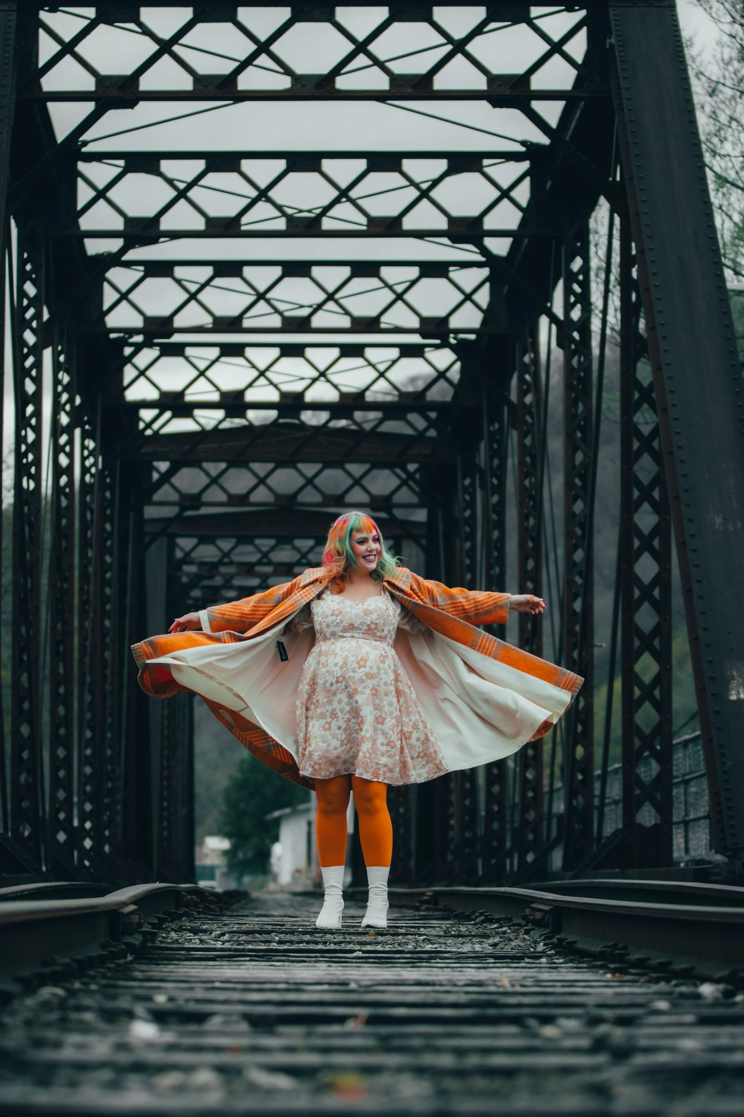 A woman with colorful hair and bright orange leggings standing on a train track under a metal bridge, wearing a floral dress and an orange checkered jacket, smiling with her arms outstretched.