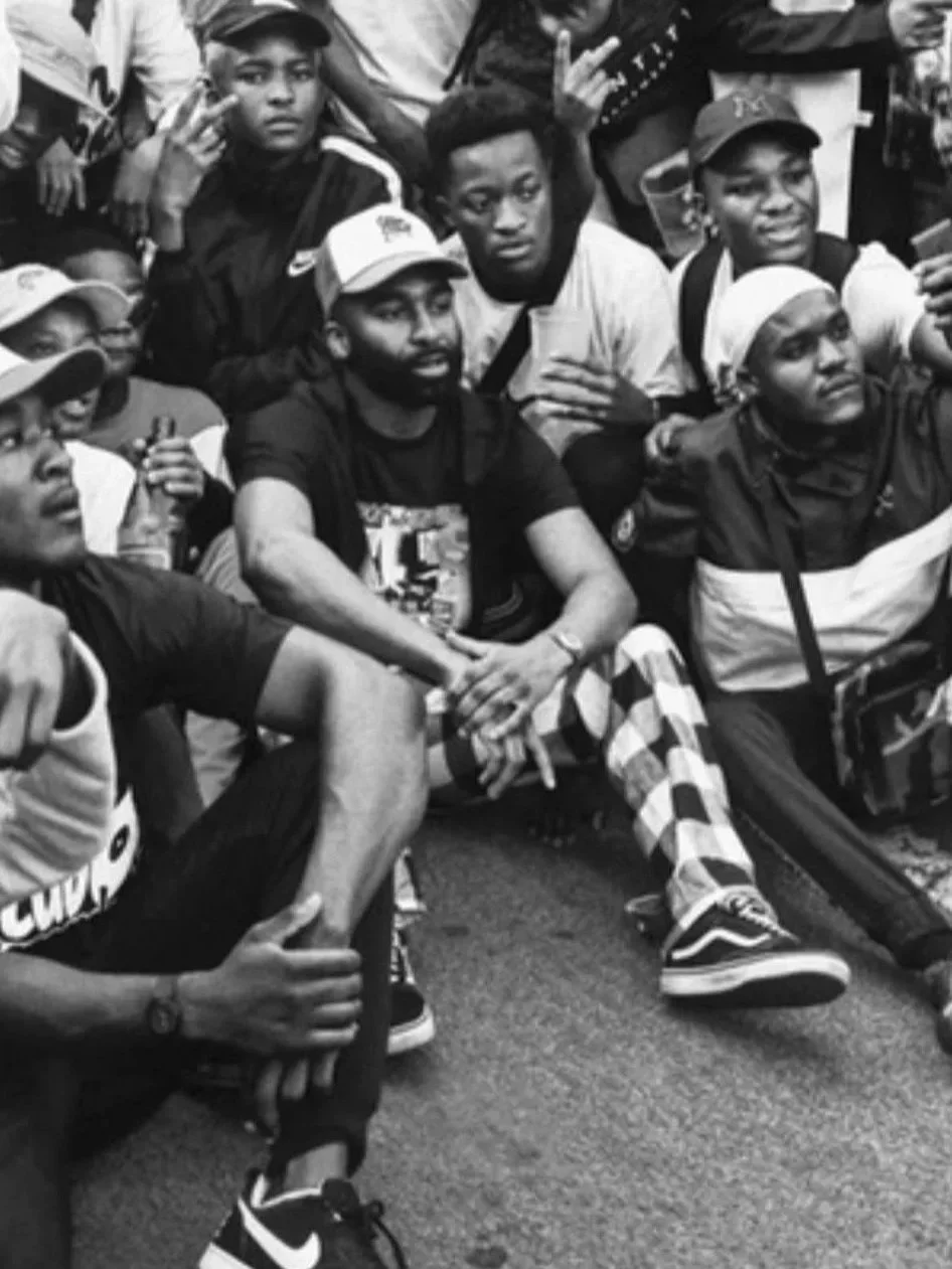 Black and white photo of the late South African rapper, Riky Rick with a group of young people gathered together.