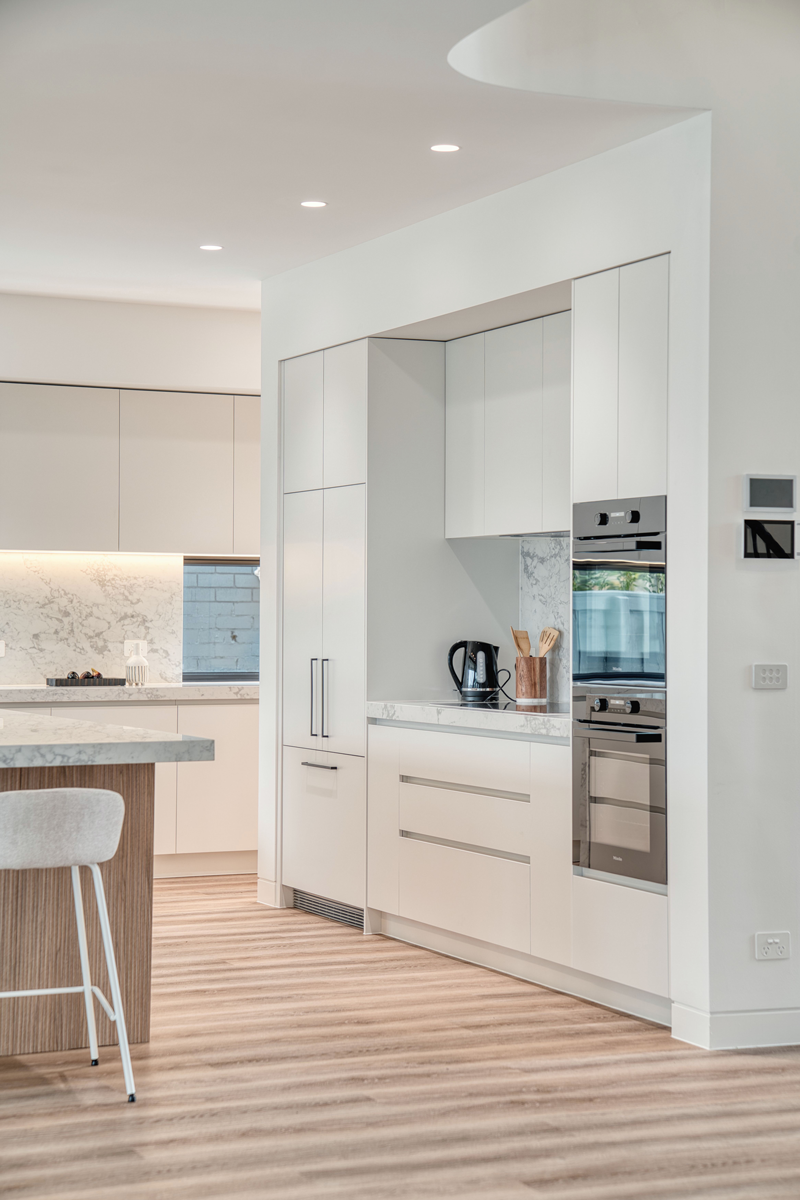 Minimalist kitchen cabinetry by Auburn Building featuring integrated Miele double ovens, a recessed coffee station with marble splashback, and seamless floor-to-ceiling white pantry storage.