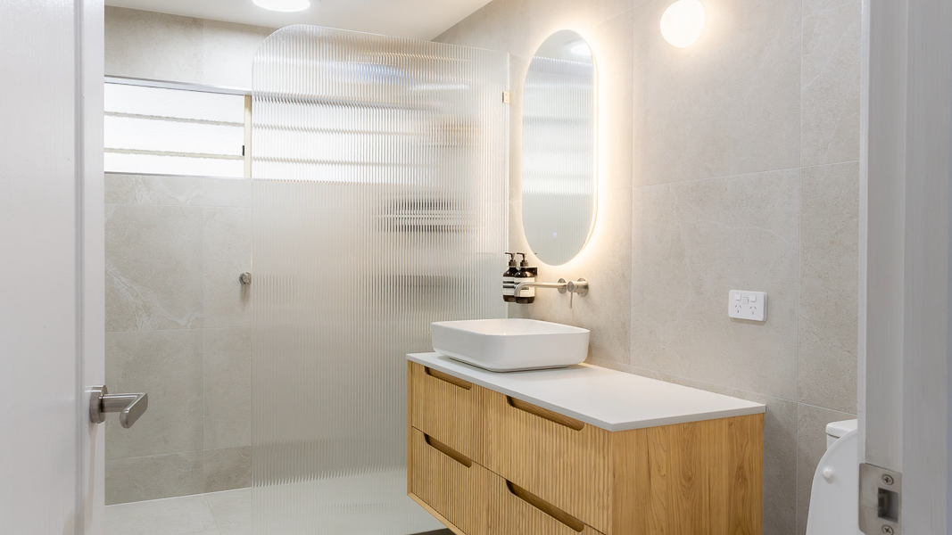 Contemporary bathroom renovation by Auburn Building featuring a fluted glass shower screen, a floating timber vanity with vertical slats, a white vessel sink, and a backlit oval mirror.