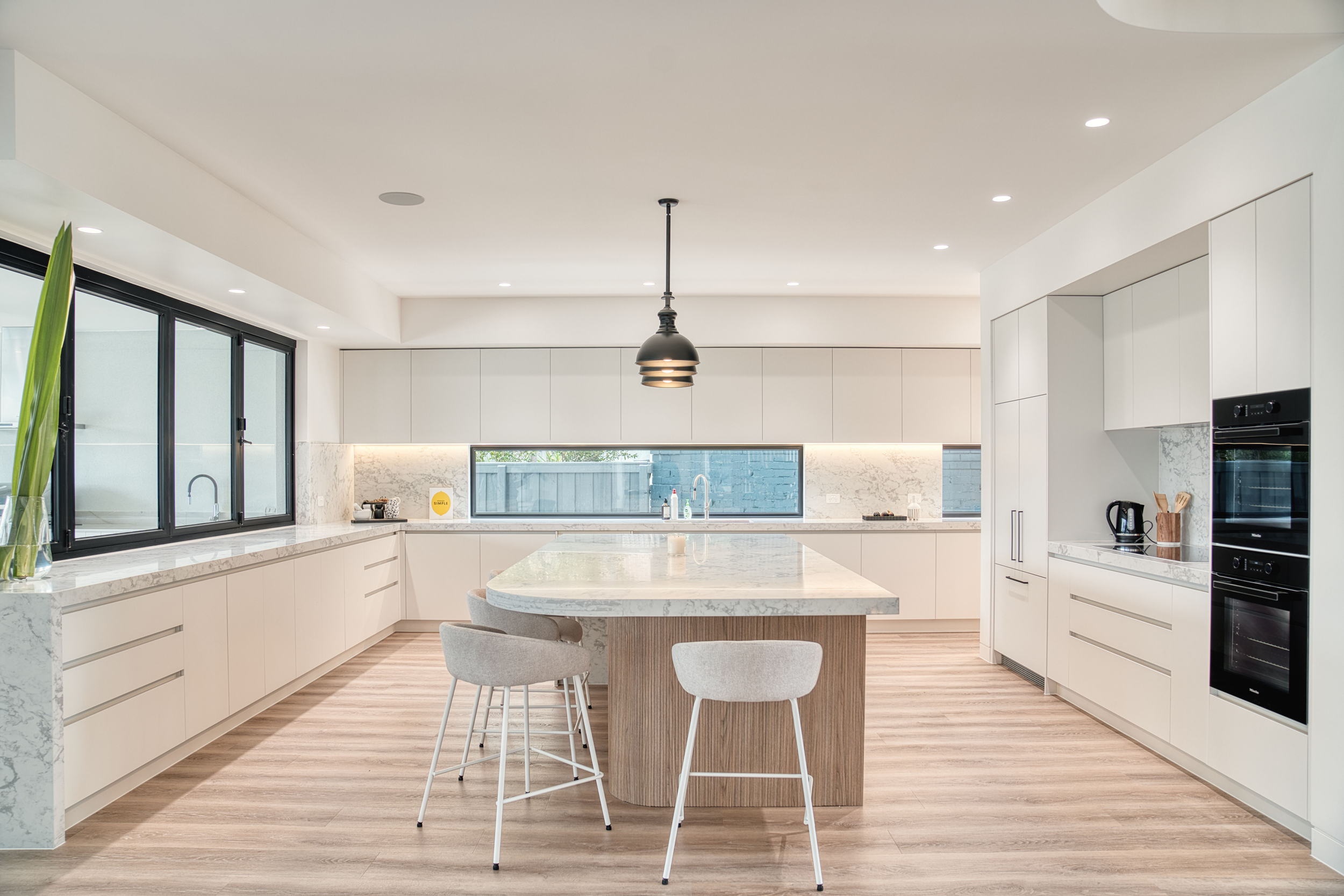 Modern custom kitchen by Auburn Building in Melbourne featuring an expansive marble-topped island with a curved wood base, white minimalist cabinetry, integrated Miele appliances, and a unique horizontal window splashback.