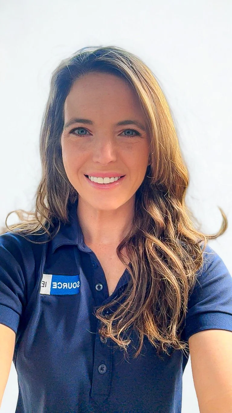 A woman with long wavy brown hair, blue eyes, and fair skin, smiling, wearing a navy blue polo shirt with a partial logo or text on the left side.