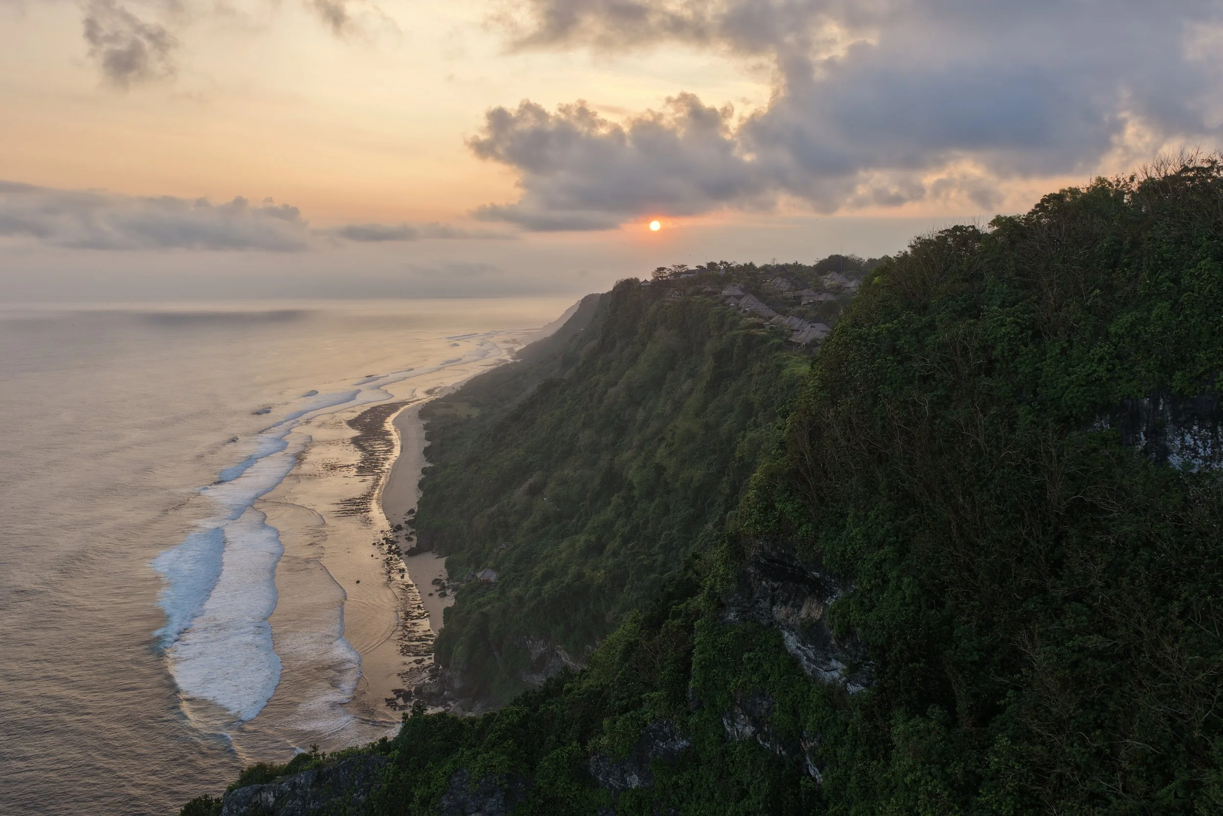 Sunset over a coastal landscape with a cliff covered in greenery, a beach, and waves crashing on the shore