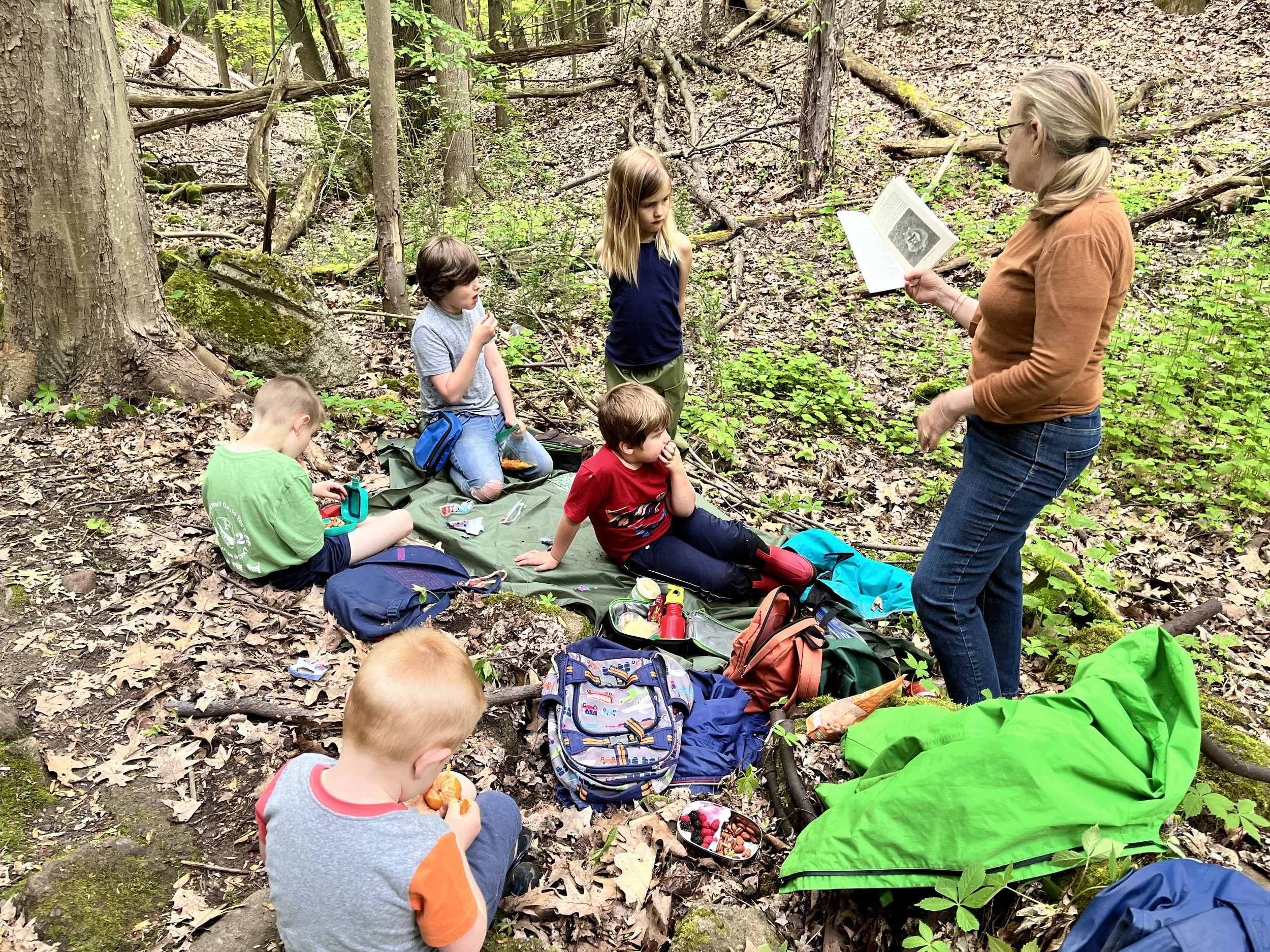 An adult woman appears to be leading an outdoor class or storytime session with five children sitting on a tarp in a forested area surrounded by trees, rocks, and foliage. The woman is holding an open book and appears to be reading to the children, who are attentively listening.
