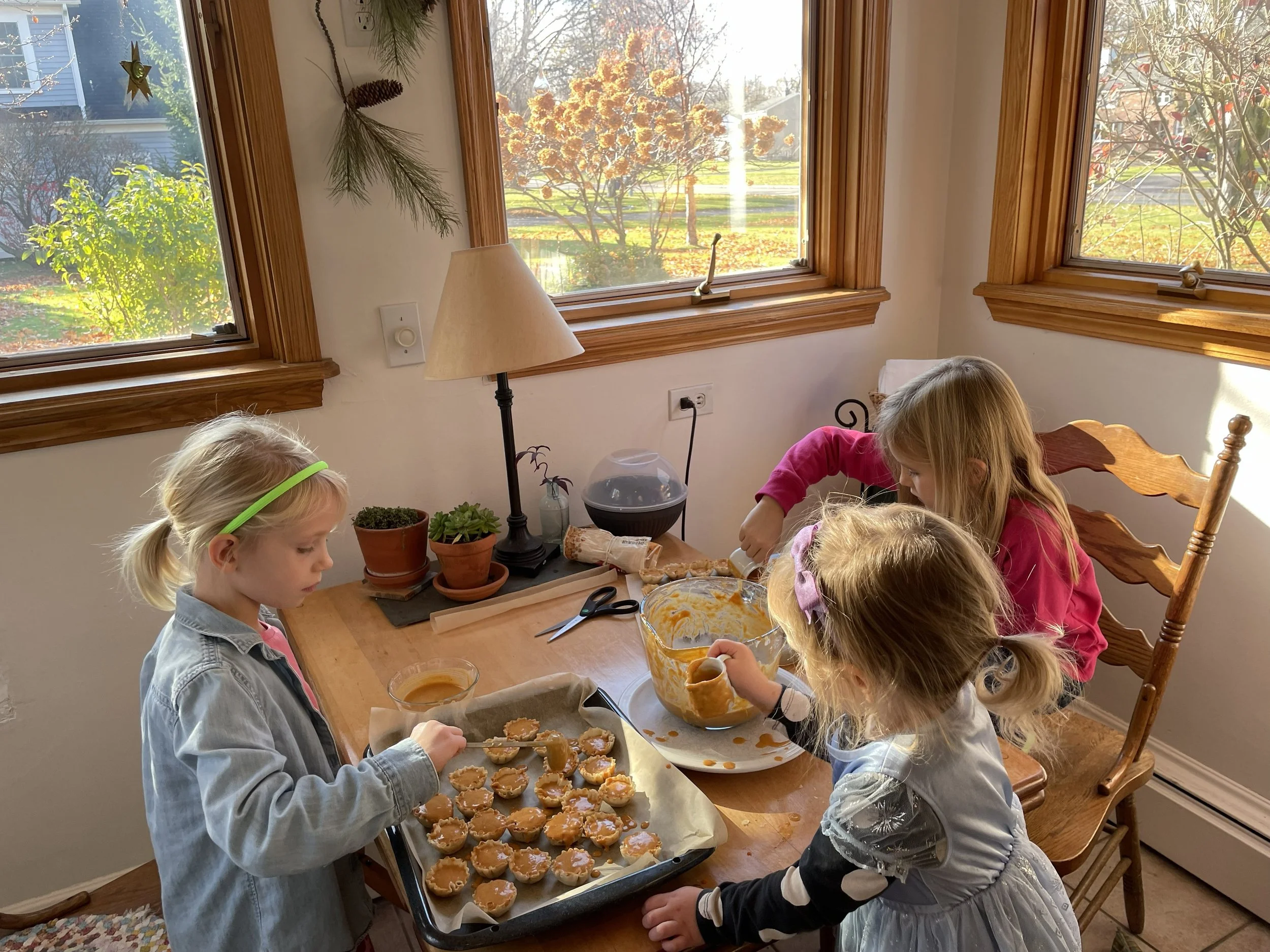 Four children preparing pumpkin-shaped cookies in a cozy kitchen with natural light. One girl is decorating cookies on a tray, while another pours orange icing into a bowl. The scene is warm and festive, with autumn leaves visible outside the window.