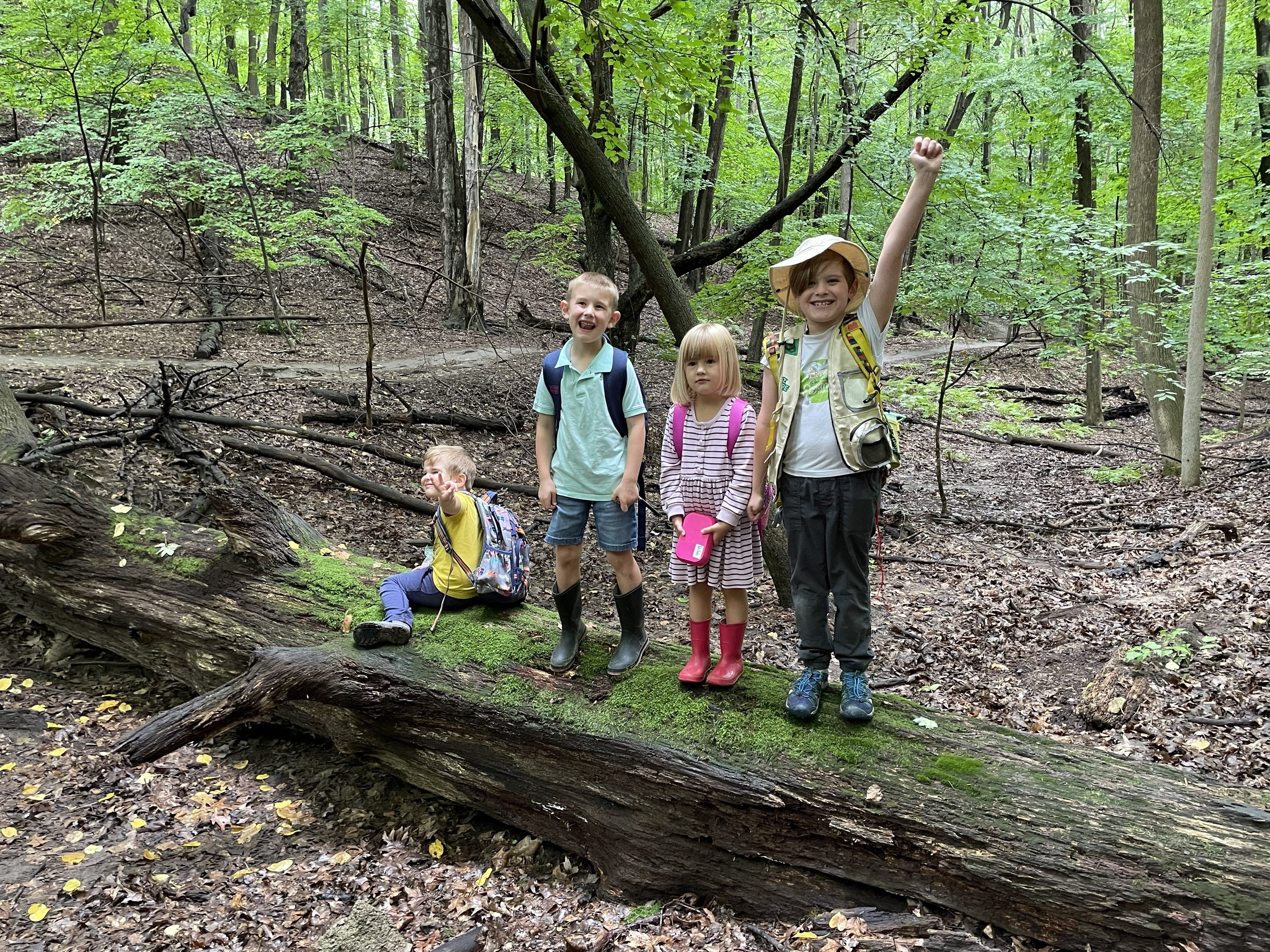 Four children standing on a fallen tree in a green forest, some smiling and one waving, wearing backpacks and rain boots.