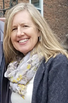A smiling woman outdoors with blonde hair, wearing a dark jacket and a light-colored scarf, standing in front of a brick wall.