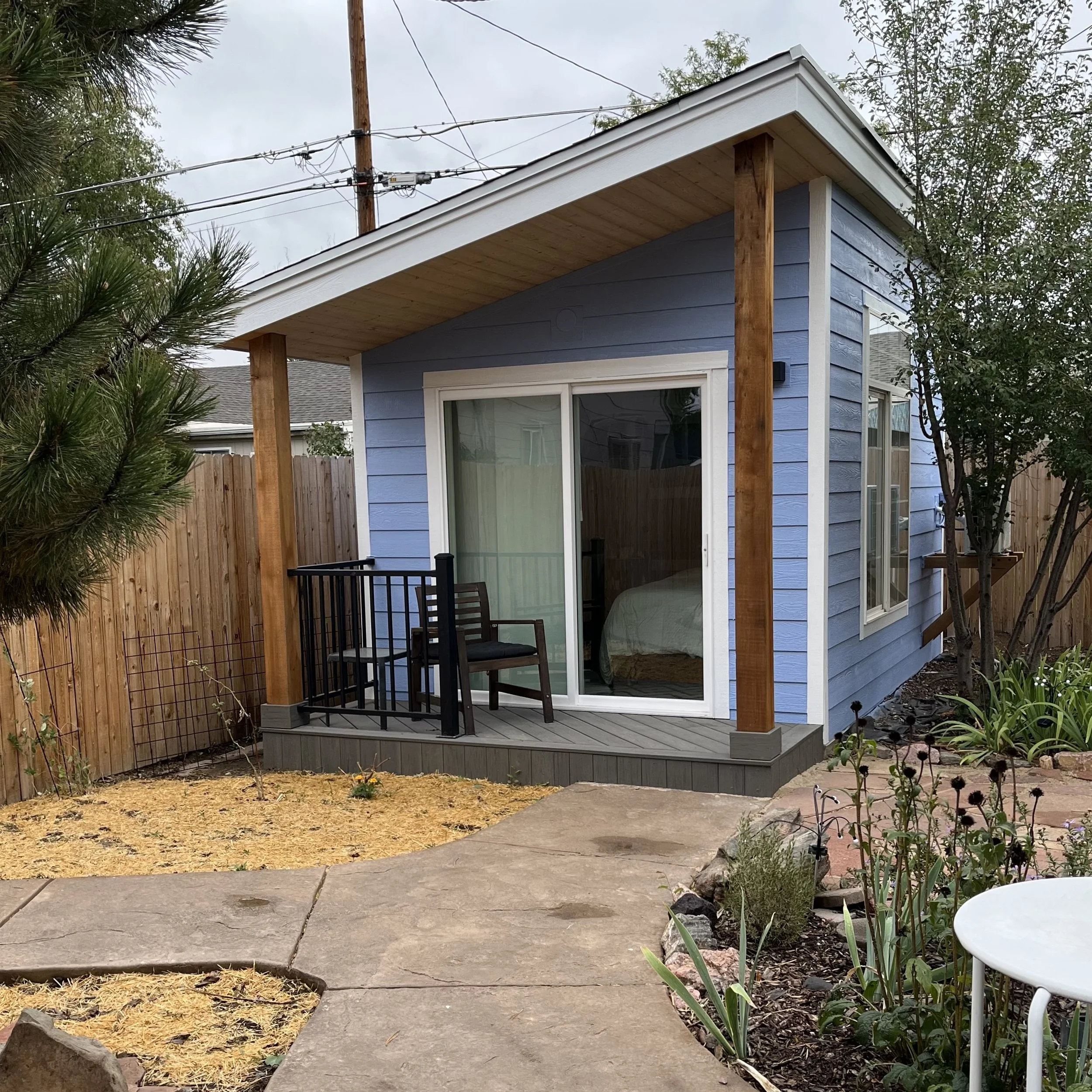 A small blue ADU with a screened porch and sliding glass door, surrounded by a yard with a concrete pathway, plants, and a wooden fence.