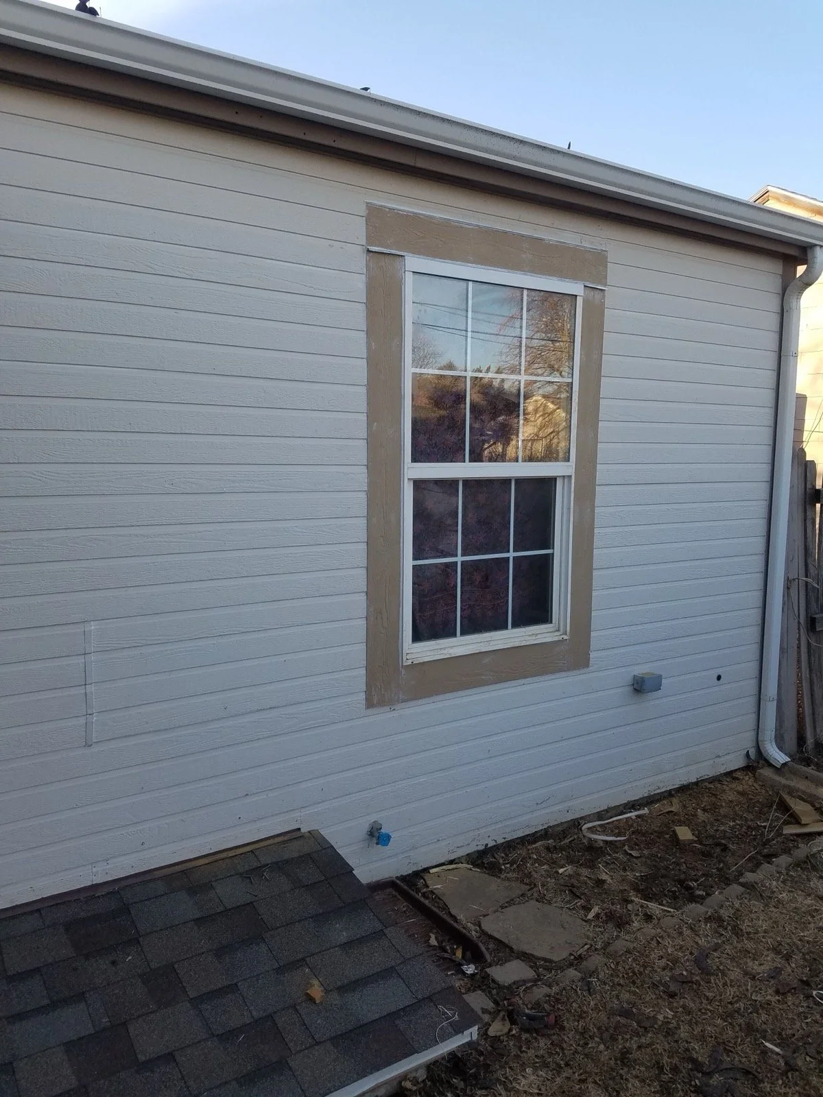 The side of a house with white siding, a newly replaced window with decorative trim, and a crawlspace hatch on the ground.