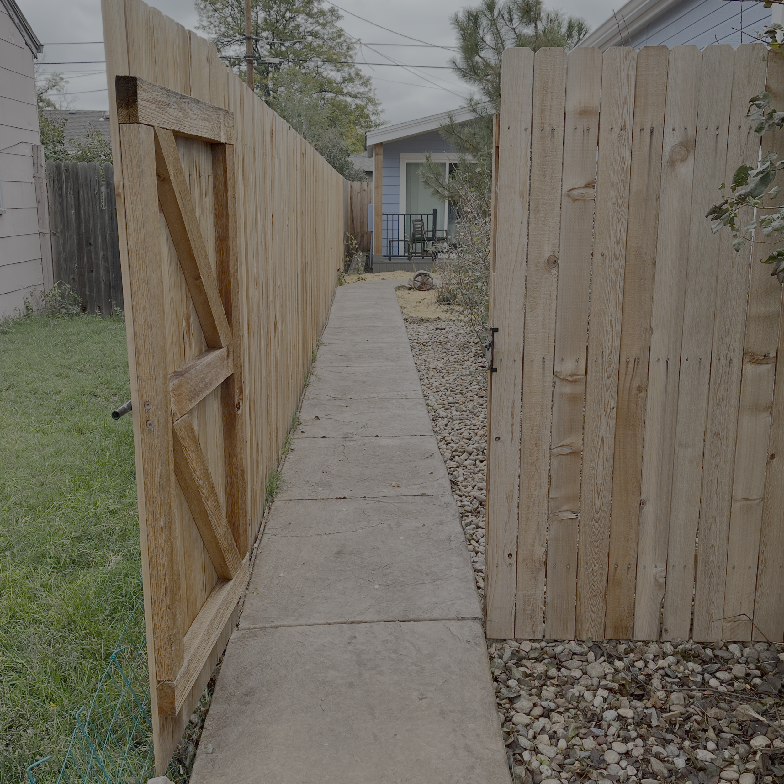 View of a residential backyard pathway with wooden fence on each side and a small gate in the left fence, leading to an ADU with a small porch and outdoor furniture in the background.