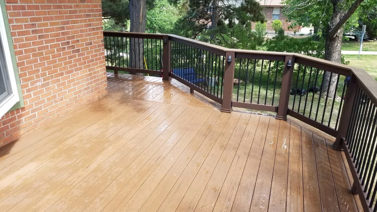 A wooden deck next to a brick wall, surrounded by a black metal railing with brown posts, overlooking a green yard with trees and some parked cars.