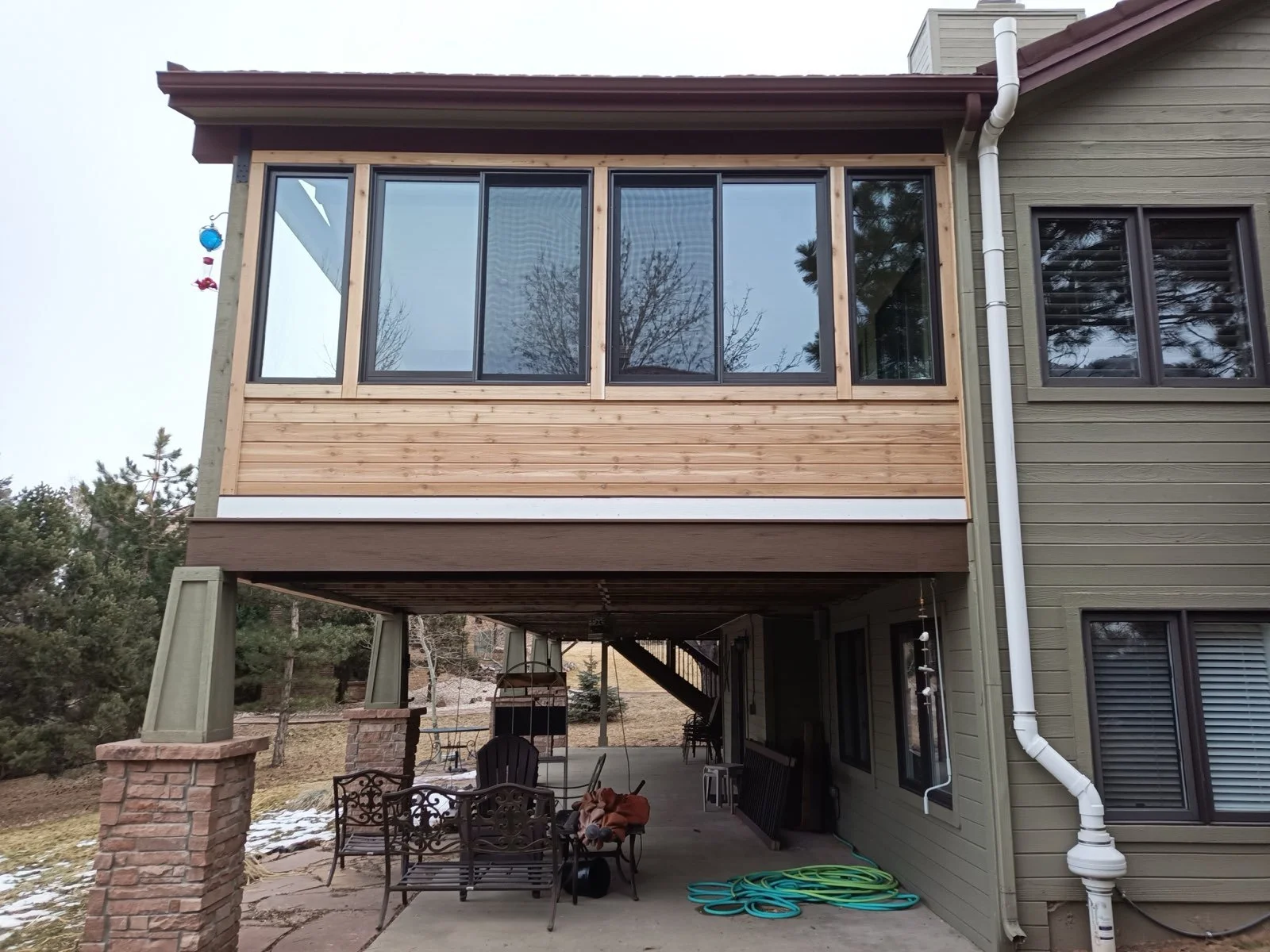 Second-story enclosed sunroom with large sliding glass doors, visible wood framing, pillows, a hanging wind chime, patio furniture, a grill, and garden hose in a backyard.