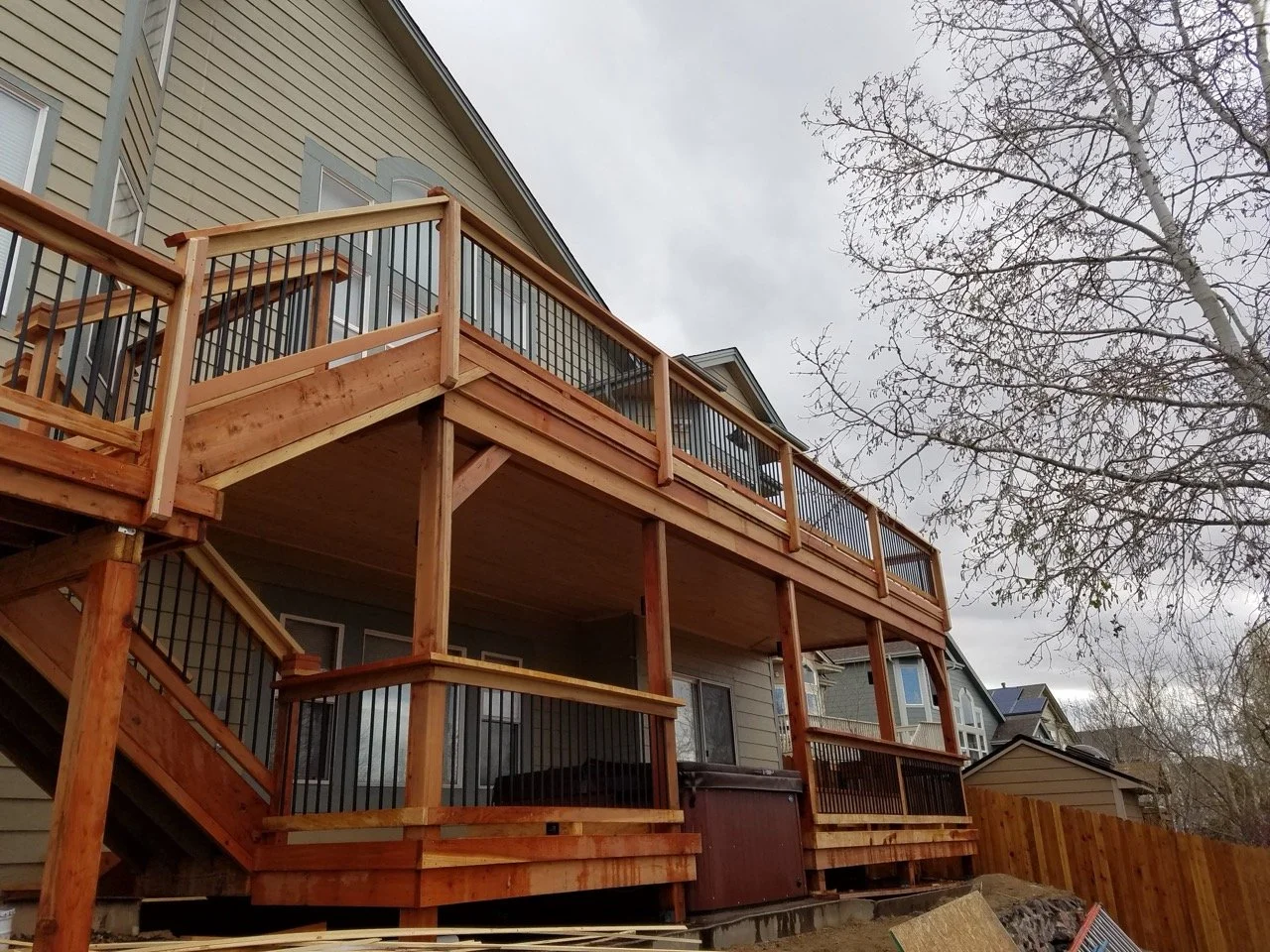 Newly built wooden deck with black metal railings on the back of a beige house, with a tree in the background and a cloudy sky.