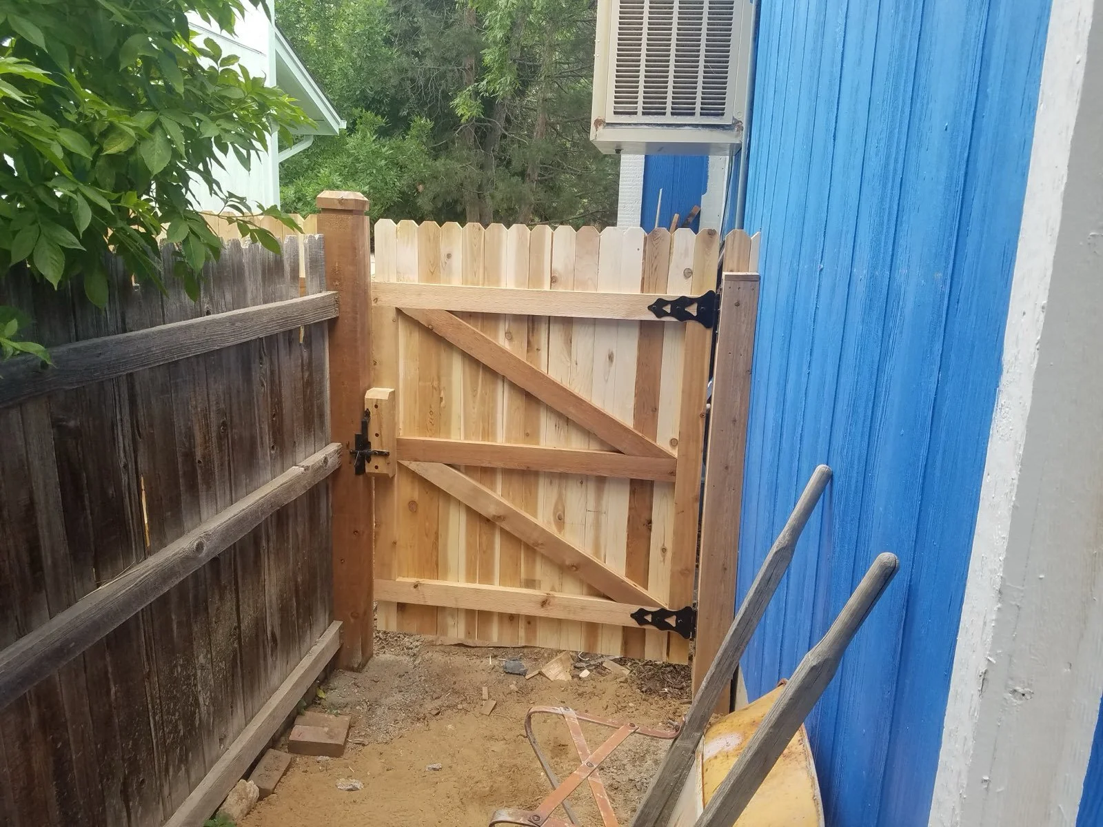 A newly installed wooden gate with black metal hardware, situated between a dark brown fence and a blue building wall, with a wheelbarrow and a ladder nearby, in a backyard with trees in the background.