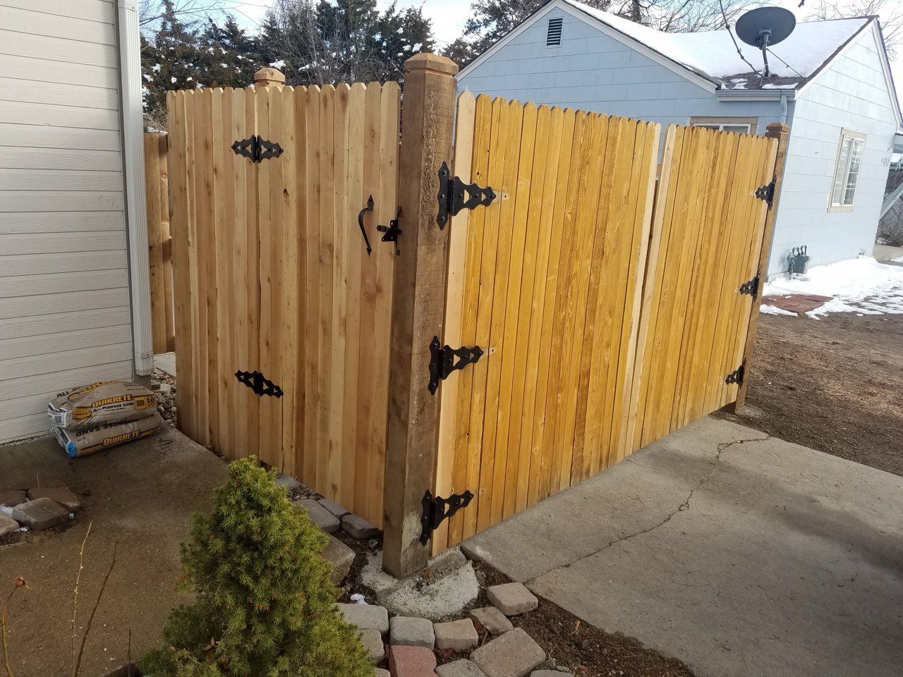 A newly installed wooden privacy fence and driveway gate with decorative black metal hinges and latch, next to a concrete driveway and house with snow on the ground.